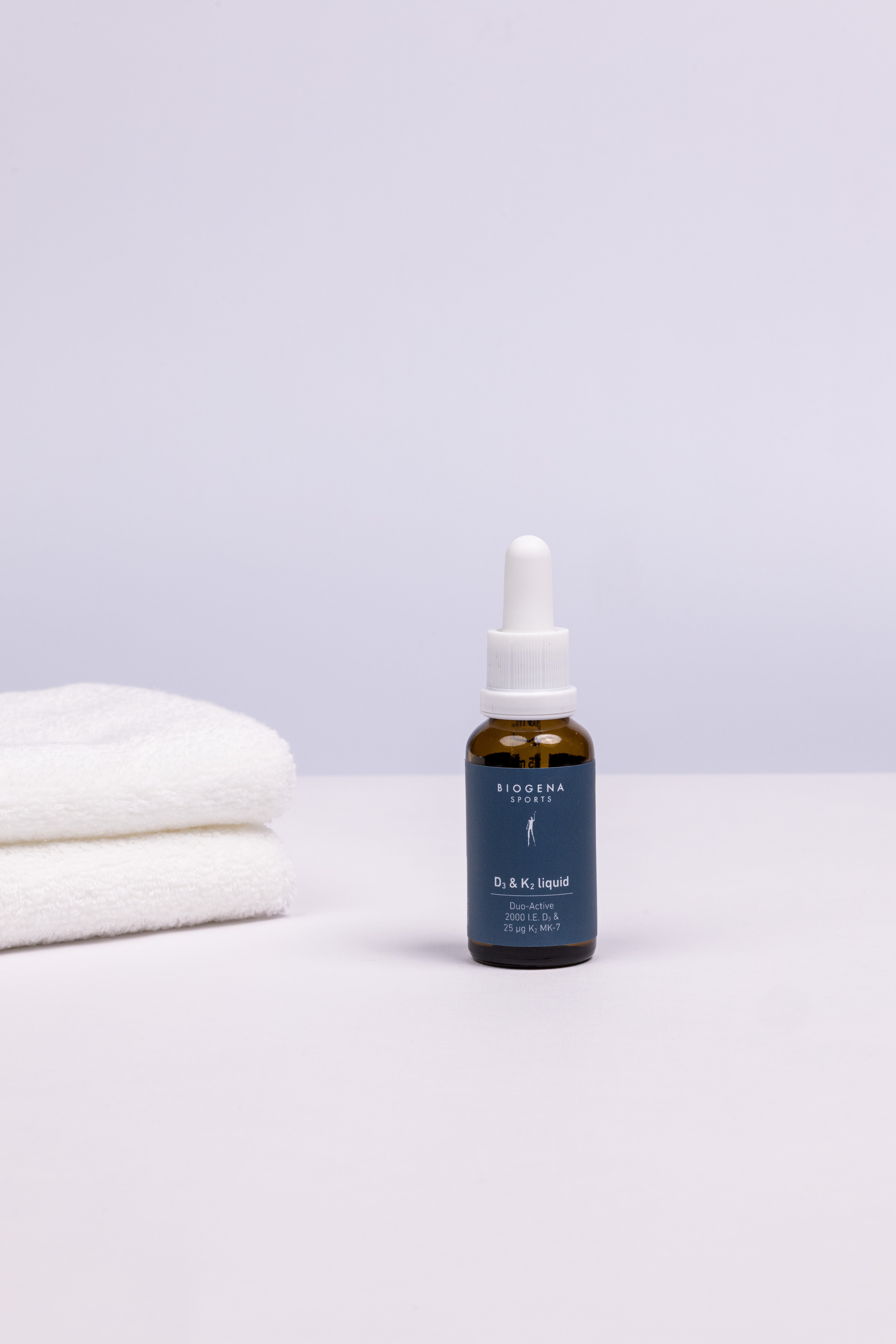 BIOGENA SPORTS D3 & K2 liquid Flasche stehend, links daneben ein Stapel weißer Handtücher.