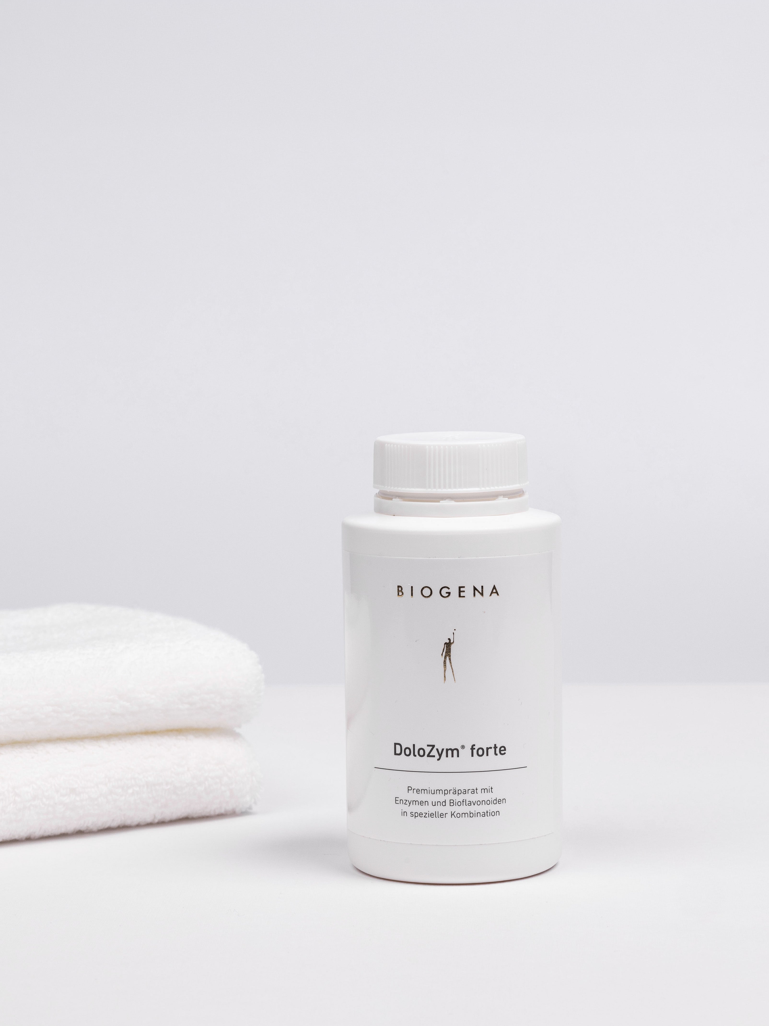 Geschlossene Verpackungsdose Biogena DoloZym forte, links daneben ein Stapel weißer Handtücher.