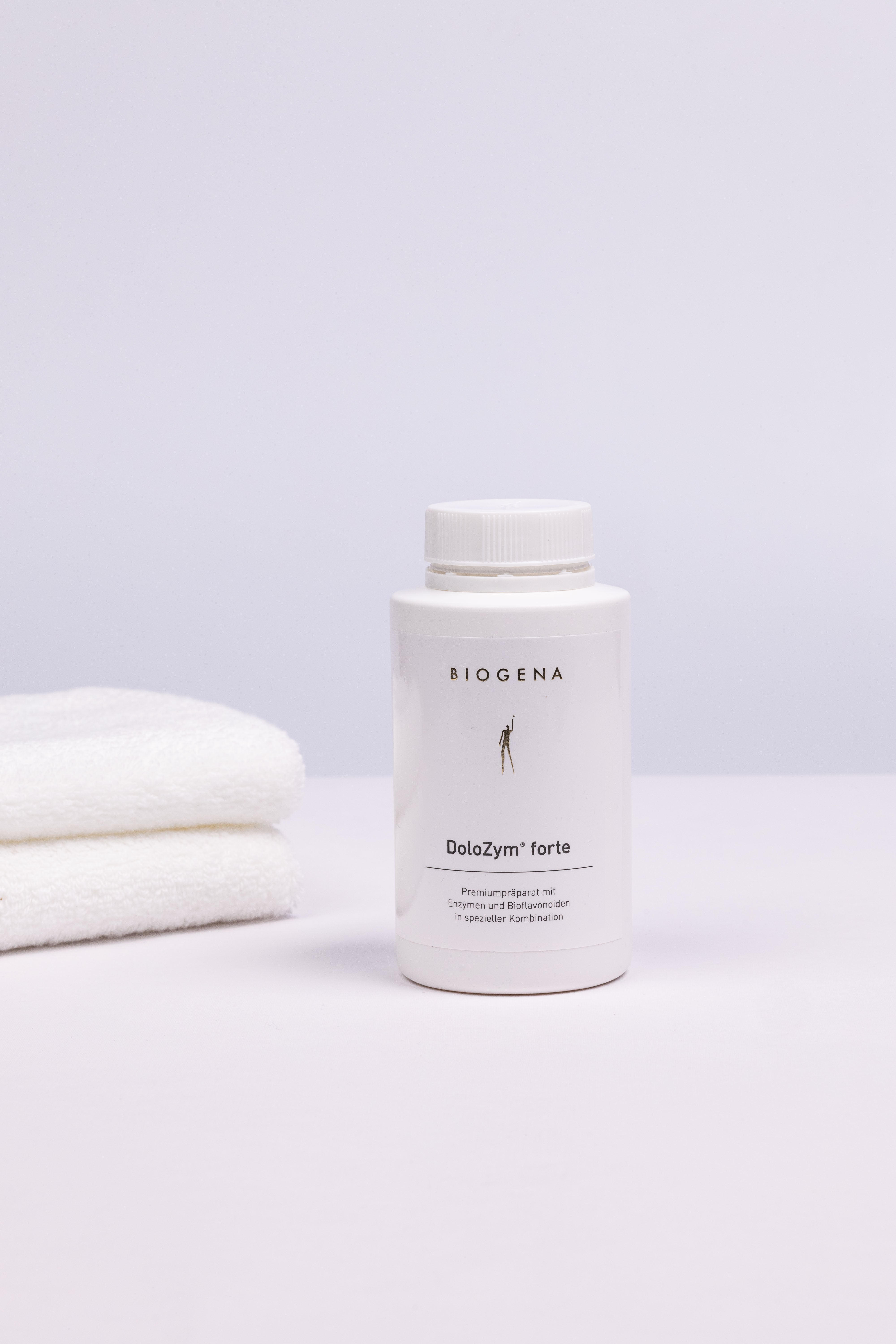 Geschlossene Verpackungsdose Biogena DoloZym forte, links daneben ein Stapel weißer Handtücher.