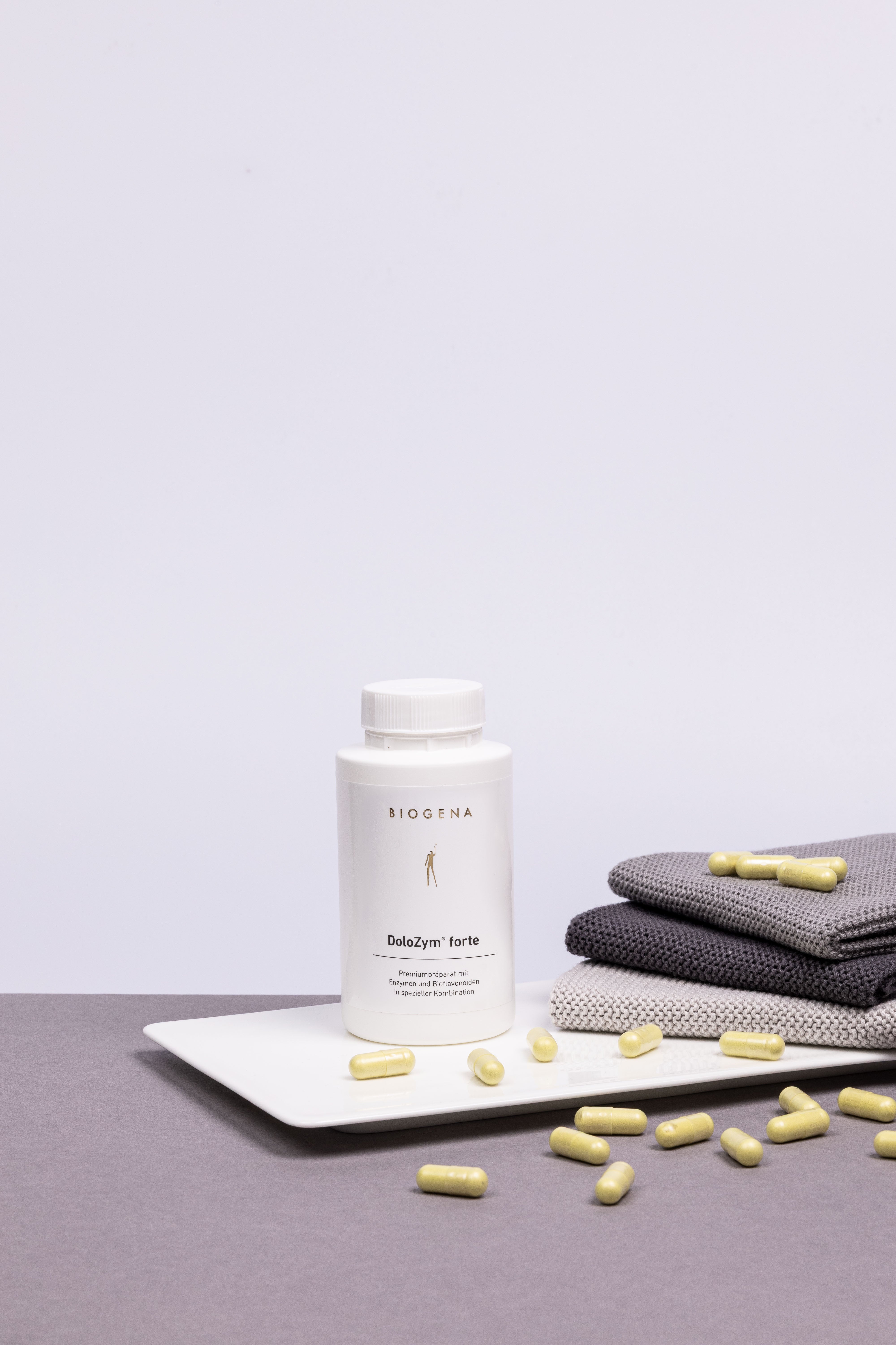 Verpackungsdose von Biogena DoloZymn forte steht auf einem weißen Tablett umgeben von gelben Kapseln, rechts daneben ist ein Stapel grauer Handtücher erkennbar.
