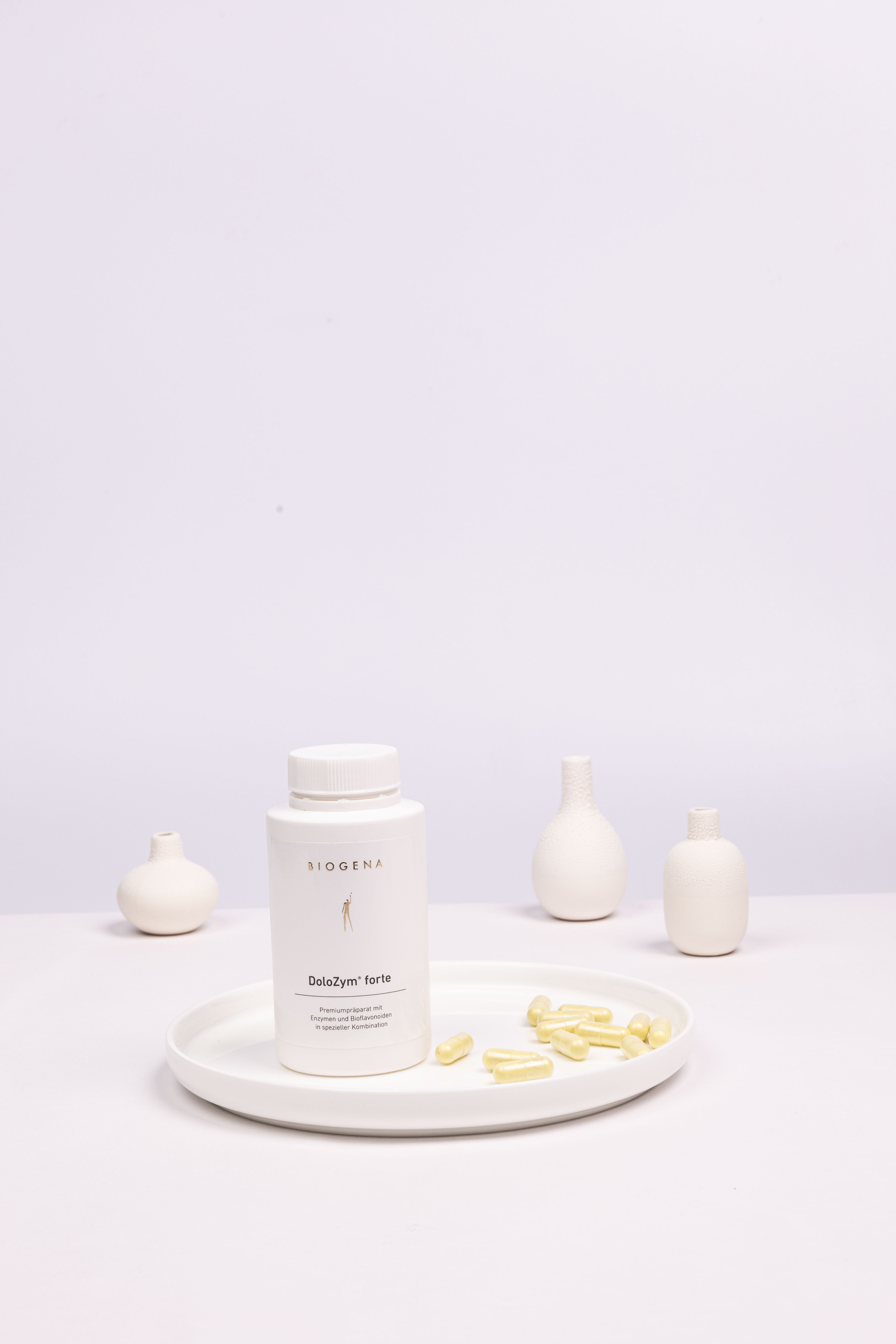 Geschlossene Verpackung von Biogena DoloZym forte steht auf einem weißen Tablett umgeben von gelben Kapseln.