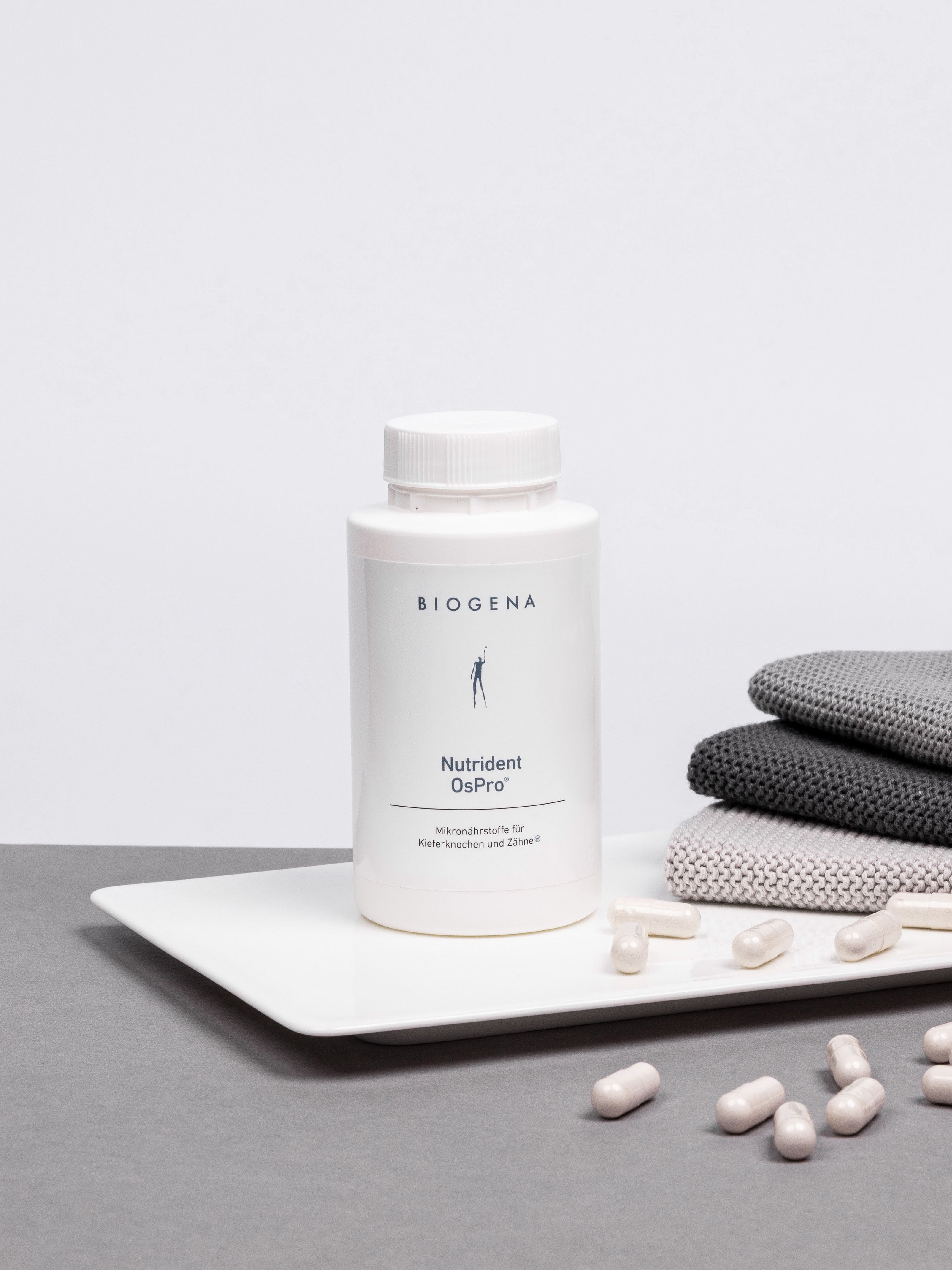 Verpackungsdose von Biogena Nutrident OsPro steht auf einem weißen Tablett umgeben von weißen Kapseln, rechts daneben ist ein Stapel grauer Handtücher erkennbar.