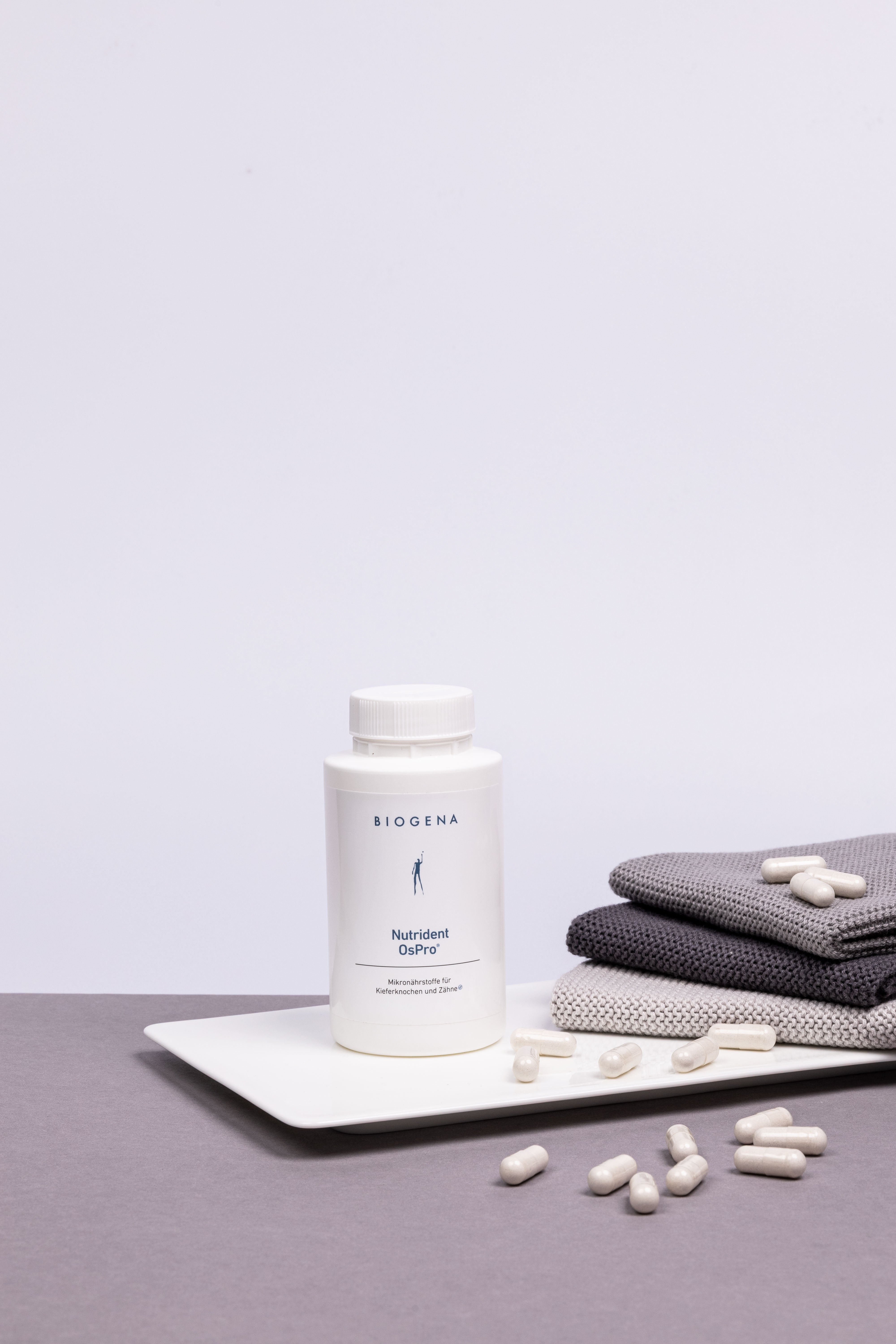 Verpackungsdose von Biogena Nutrident OsPro steht auf einem weißen Tablett umgeben von weißen Kapseln, rechts daneben ist ein Stapel grauer Handtücher erkennbar.