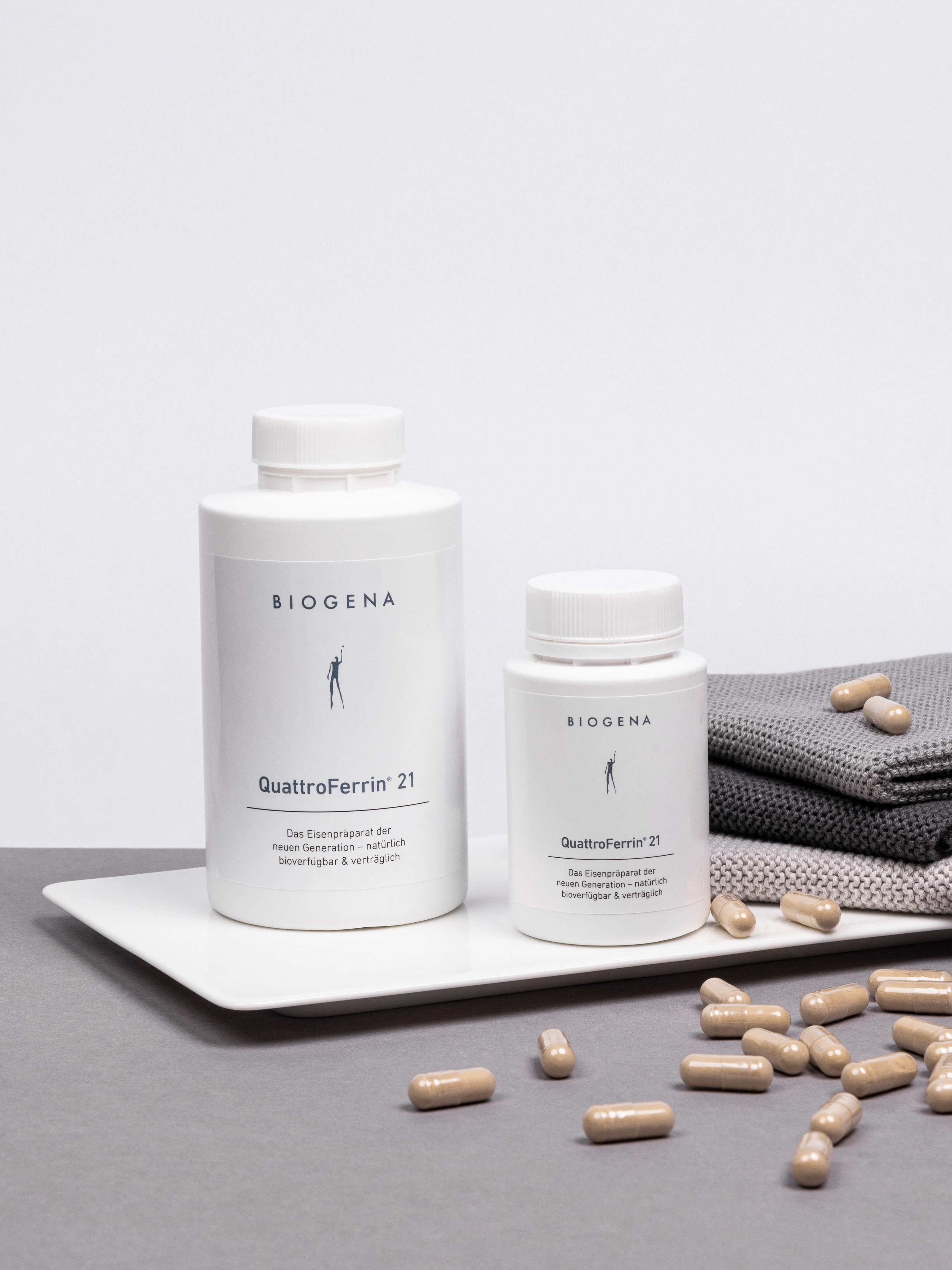 Zwei Dosen QuattroFerrin 21 stehen auf einem Tablett umgeben von Kapseln, rechts daneben ist ein Stapel grauer Handtücher erkennbar.