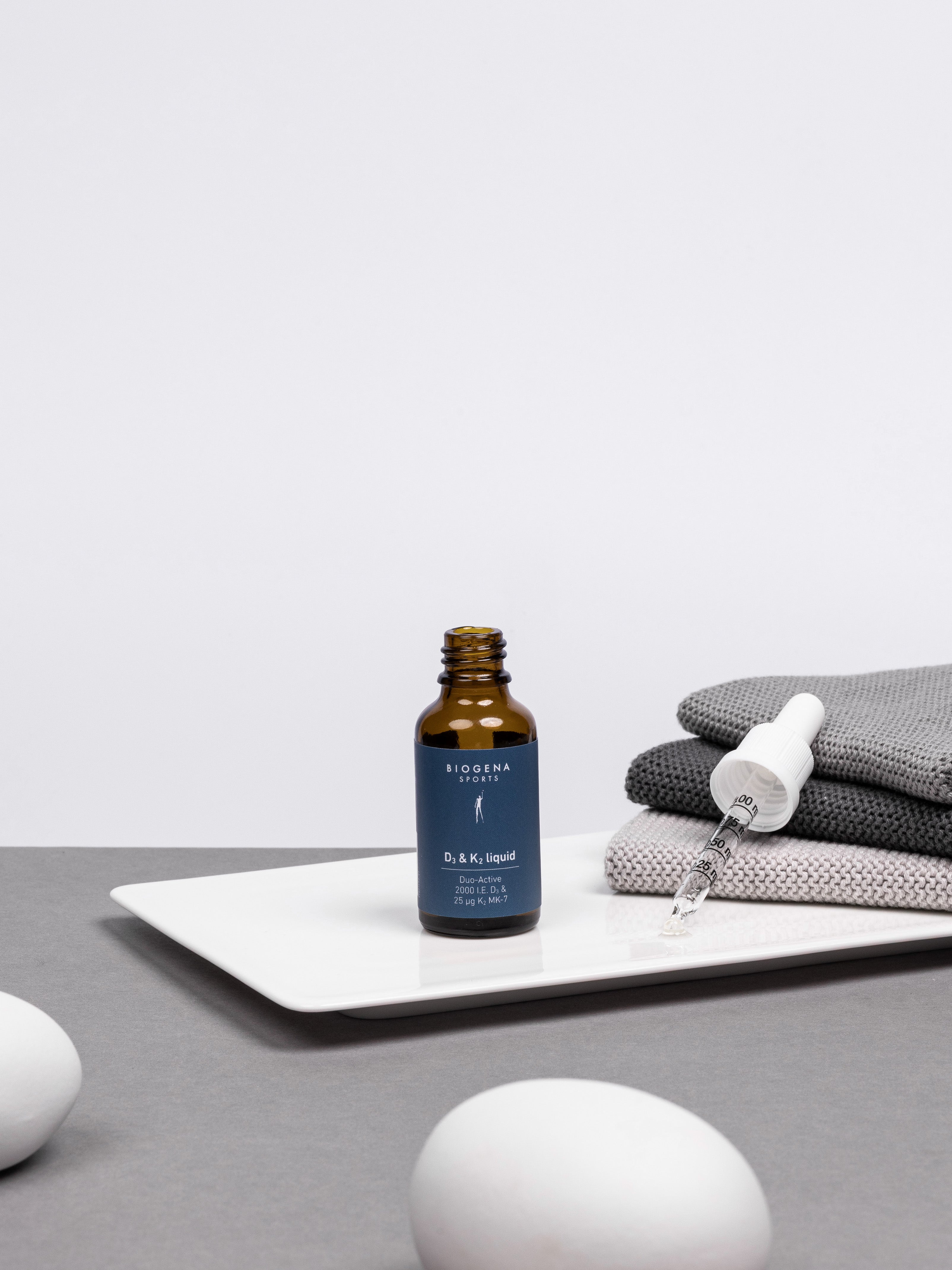 Geöffnete Flasche BIOGENA SPORTS D3 & K2 liquid Flasche stehend, daneben der Flaschendeckel mit Pipette auf einem Stapel grauer Handtücher liegend.