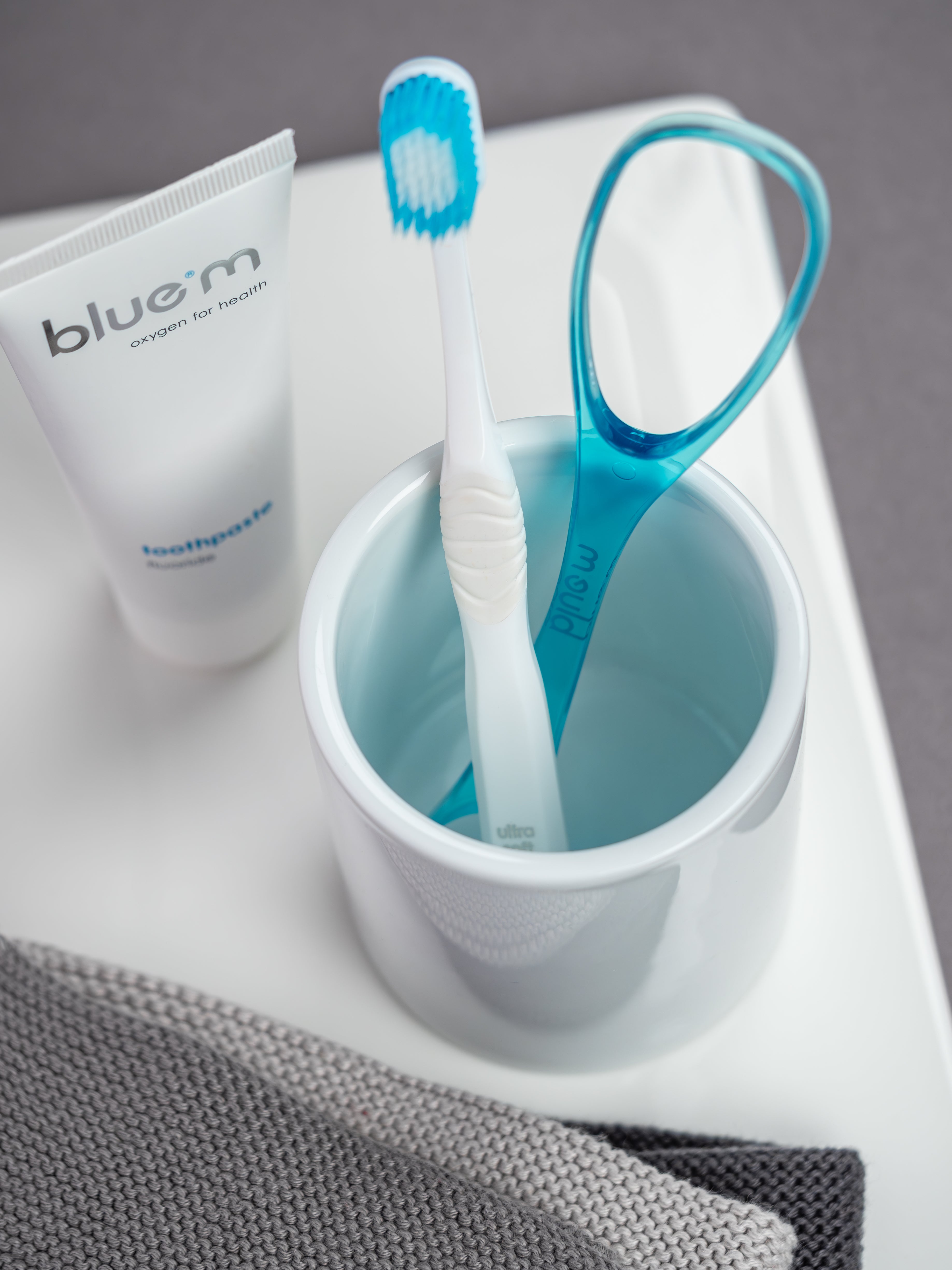 Weißer Zahnputzbecher in dem sich eine weiße Sensitiv-Zahnbürste und ein blauer Zungenreiniger befindet. Links daneben ist eine Tube Zahngel erkennbar.