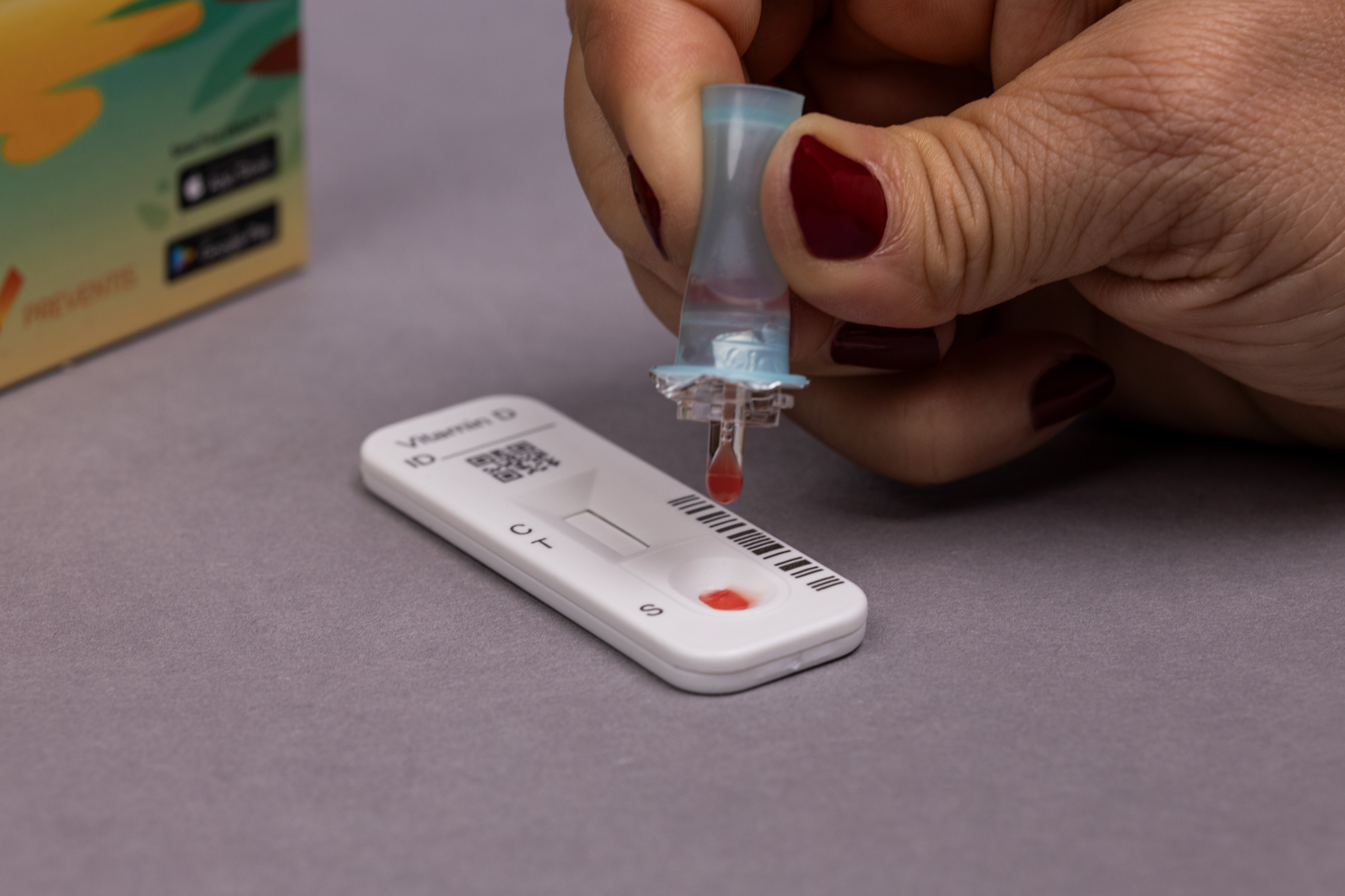 Testvorgang des Vitamin-D Gehalts, dabei hält eine Frauenhand das Probendöschen mit Pufferlösung und Blut in den Händen und tropft einen Tropfen der Lösung auf die Testkassette.