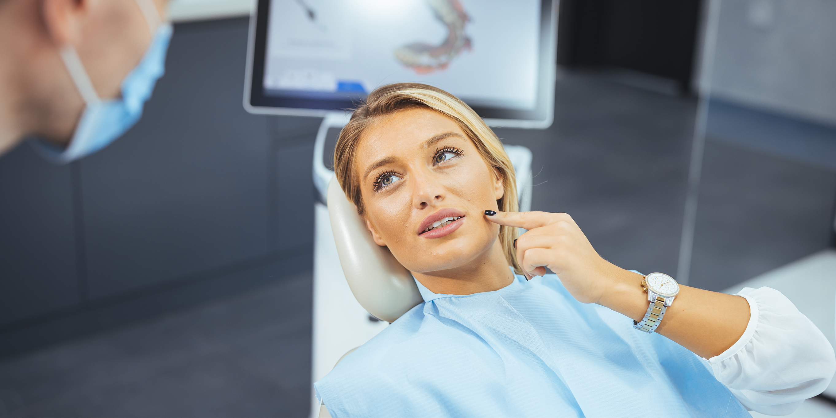 Junge Patientin liegt im Behandlungsstuhl und deutet mit ihrem linken Zeigefinger auf ihren Oberkiefer. Ein Zahnarzt sitzt leicht zu ihr vorgebeugt links neben ihr.
