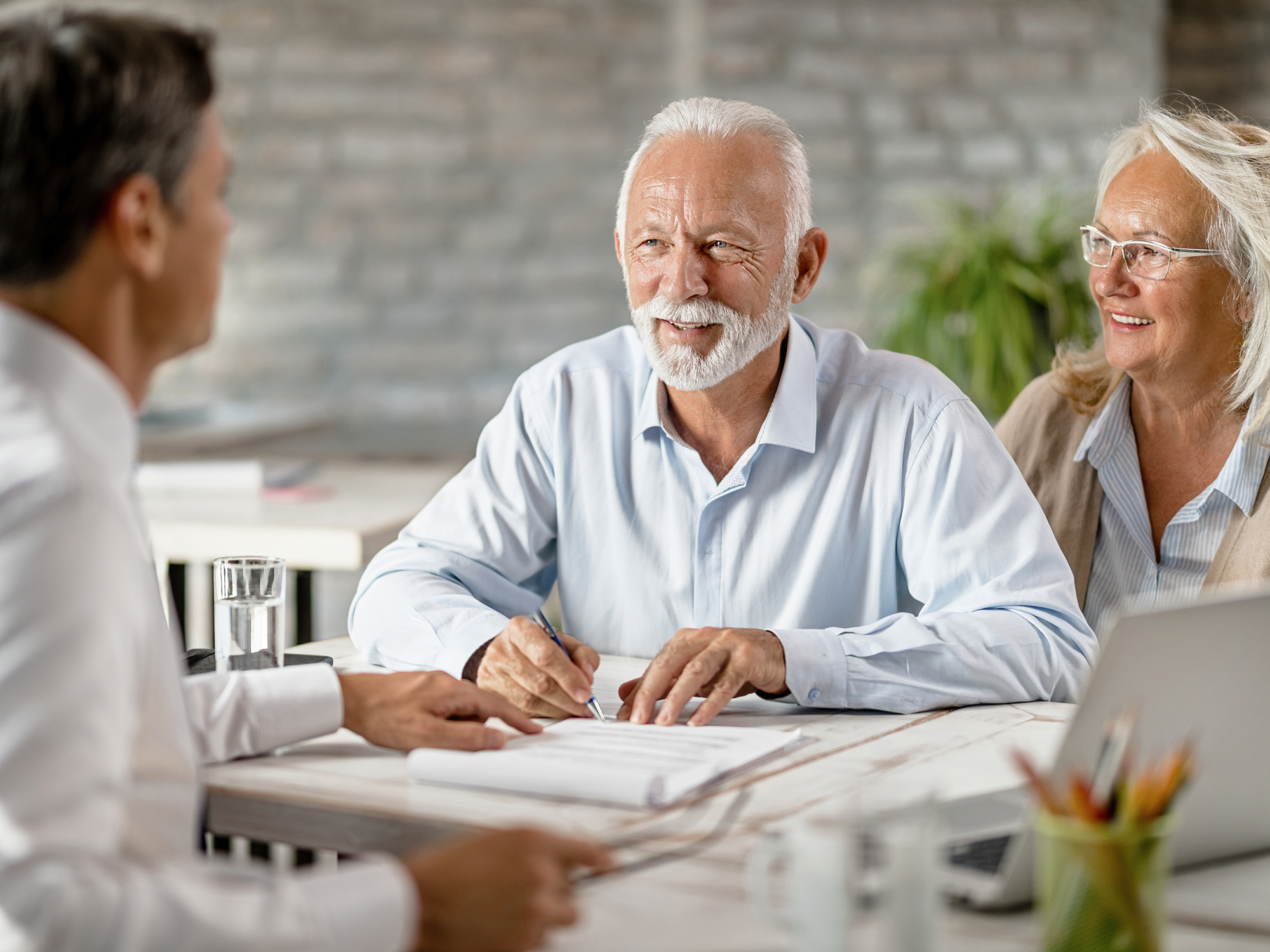 Älterer Mann und seine Frau sitzen im Behandlungszimmer am Schreibtisch des Zahnarztes und werden von dem Zahnarzt beraten.