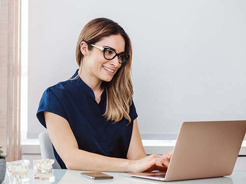 Junge Frau mit blonden Haaren und einer schwarzen Brille sitzt am Laptop.