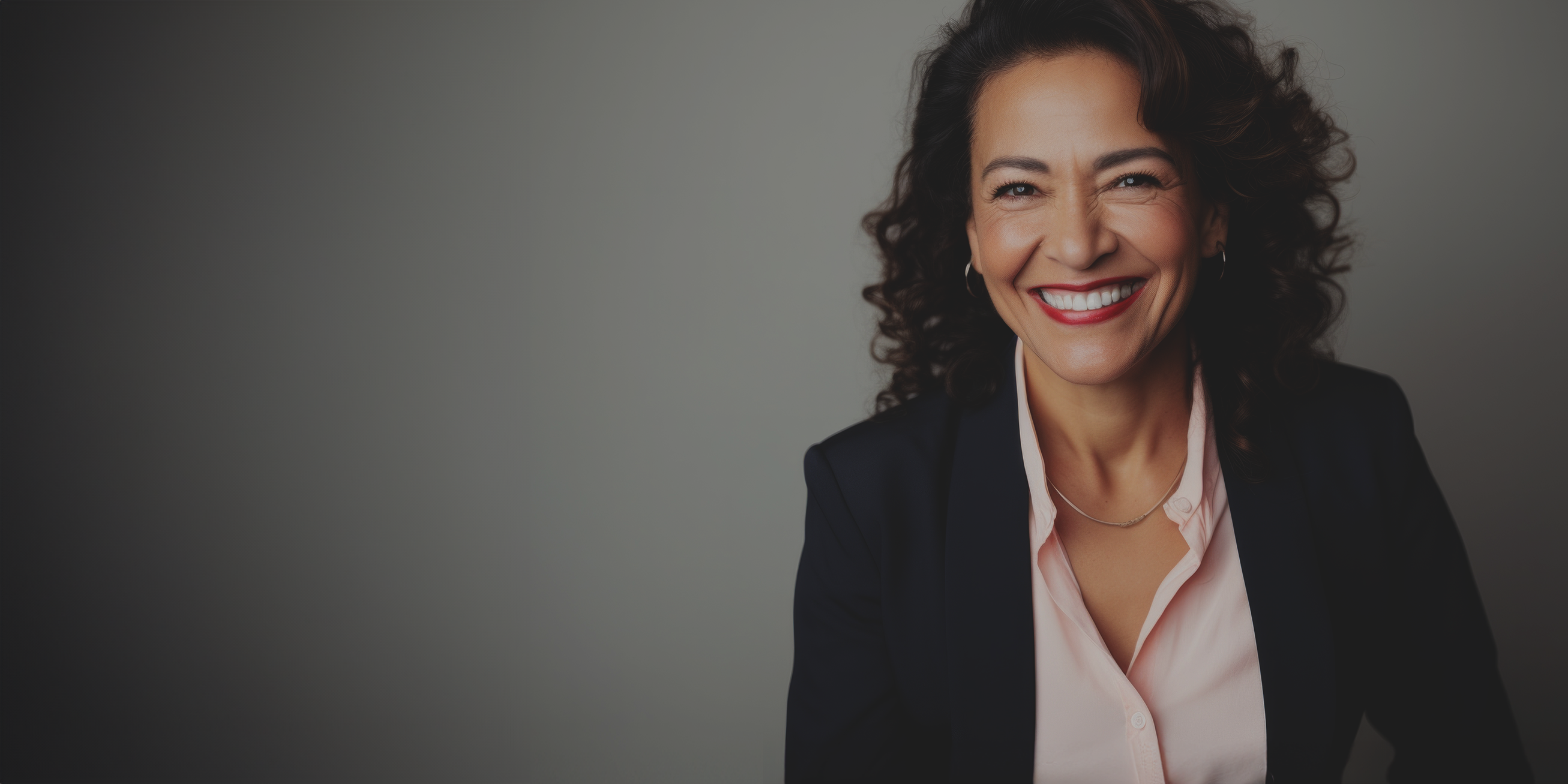 Frau mittleren Alters mit dunklen lockigen Haaren trägt einen Businessanzug und lächelt in die Kamera.