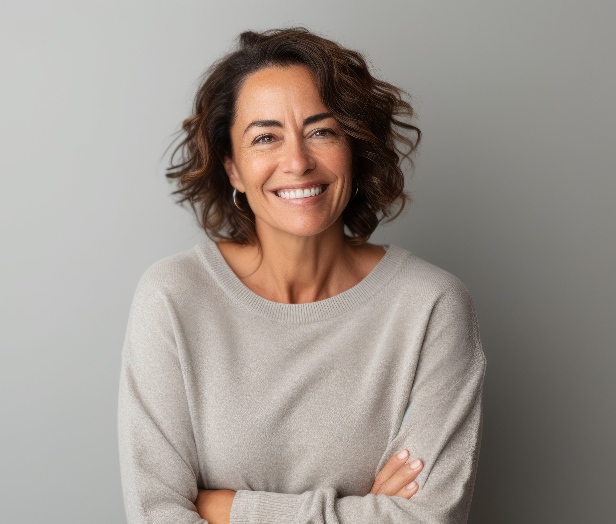 Frau mittleren Alters mit halblangen braunen Haaren hat die Arme verschränkt und lächelt in die Kamera.