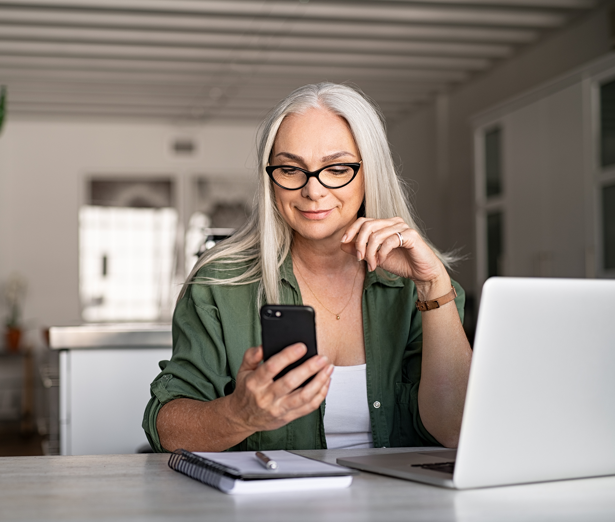 Ältere Frau mit langen weißen Haaren sitzt am Tisch mit dem Laptop vor ihr. In der rechten Hand hält sie ein Smartphone zu welchem sie lächelnd hinschaut.