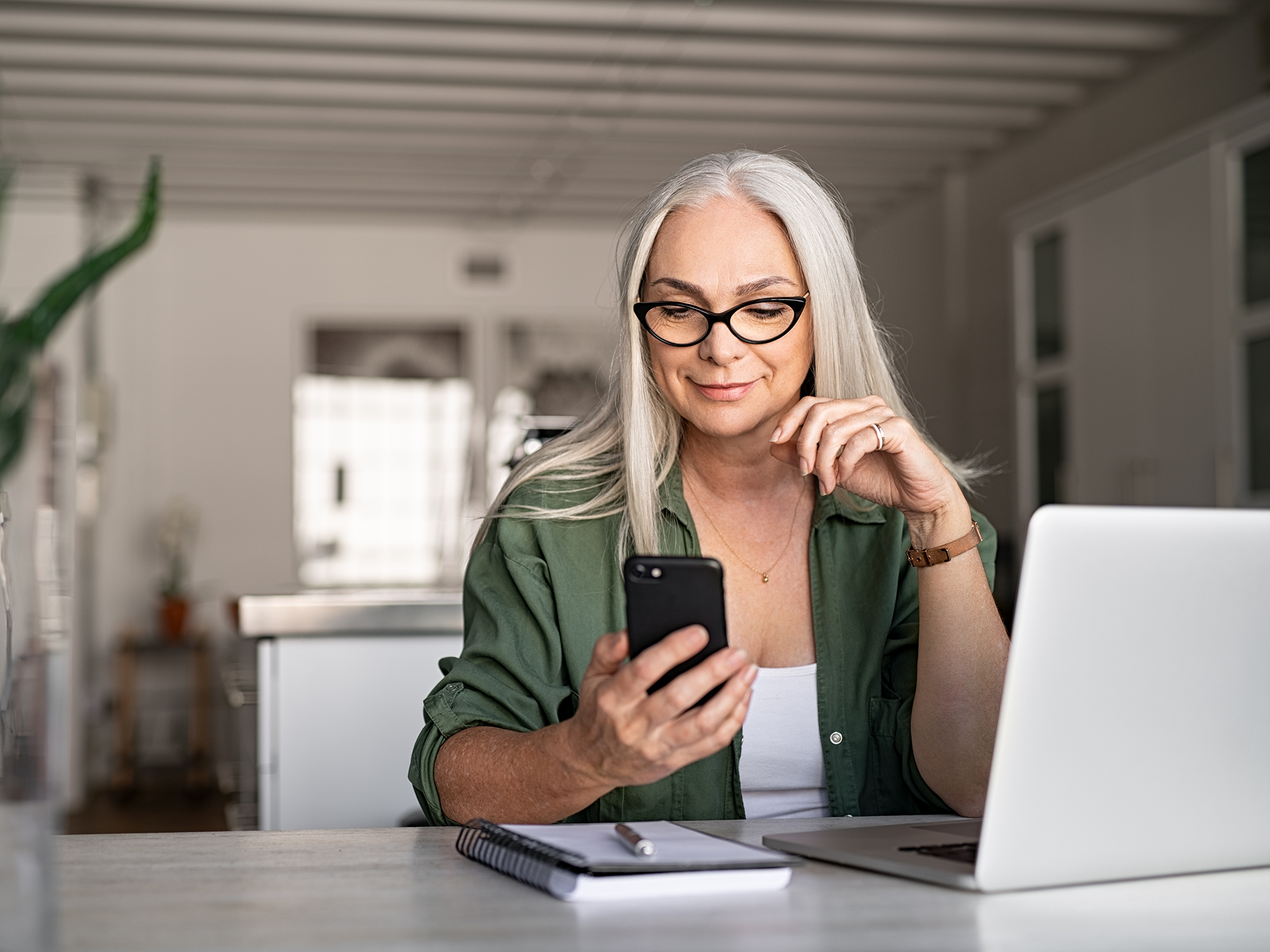 Ältere Frau mit langen weißen Haaren sitzt am Tisch mit dem Laptop vor ihr. In der rechten Hand hält sie ein Smartphone zu welchem sie lächelnd hinschaut.