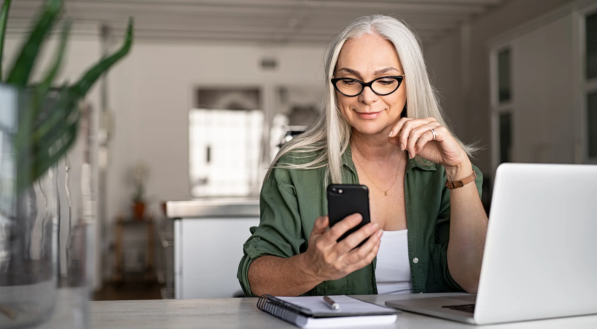 Ältere Frau mit langen weißen Haaren und einer schwarzen markanten Brille sitzt am Tisch mit einem Laptop und schaut in ein Smartphone.