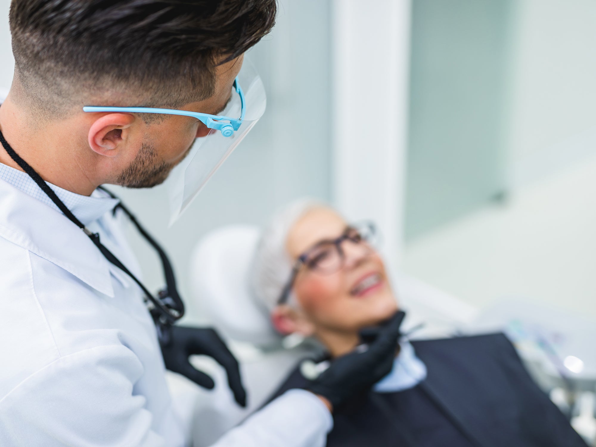 Zahnarzt mit Patientin, die lächelnd auf dem Behandlungsstuhl liegt. Der Zahnarzt trägt einen Augen-Mund-Schutz.