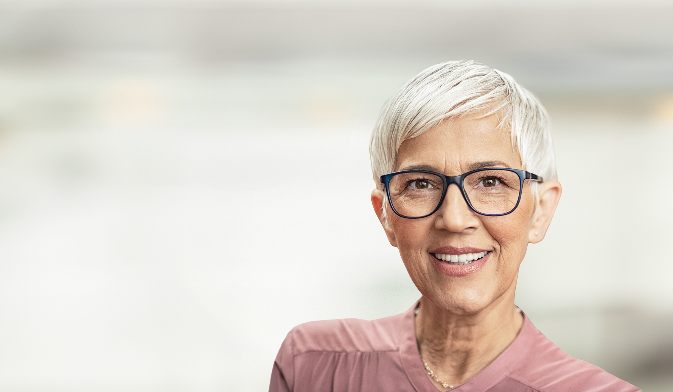 Ältere Frau mit weißen, kurzen Haaren und einer markanten schwarzen Brille, schaut zufrieden und lächelt.