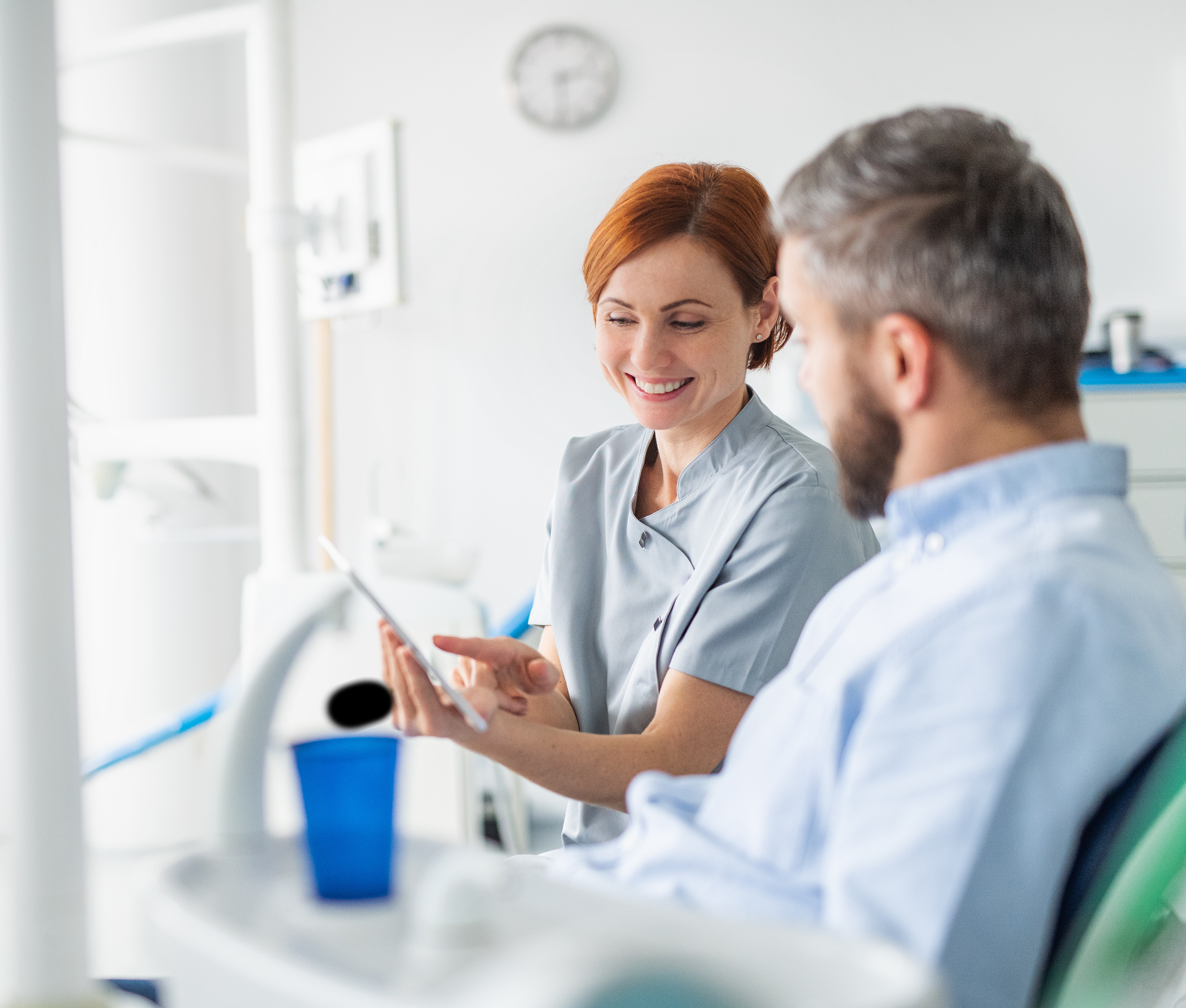 Zahnärztin hält ein Tablett in der Hand und vermittelt dem Patienten auf dem Behandlungsstuhl Informationen.