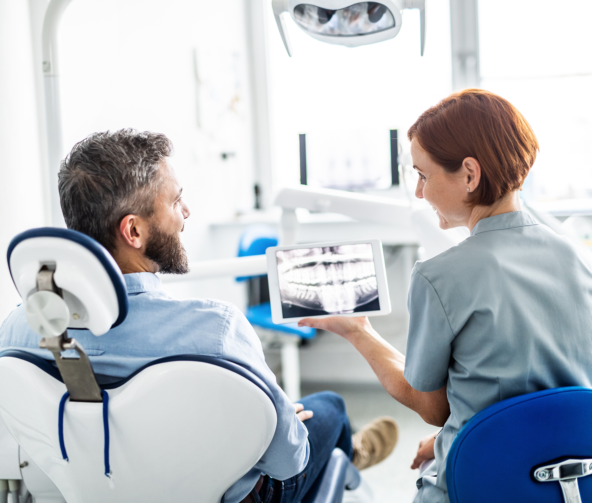 Junge Zahnärztin zeigt ihrem Patienten mit einem Tablet eine Röntgenaufnahme seines Ober- und Unterkiefers. Der Patient befindet sich dem Behandlungsstuhl, rechts daneben die junge Zahnärztin.