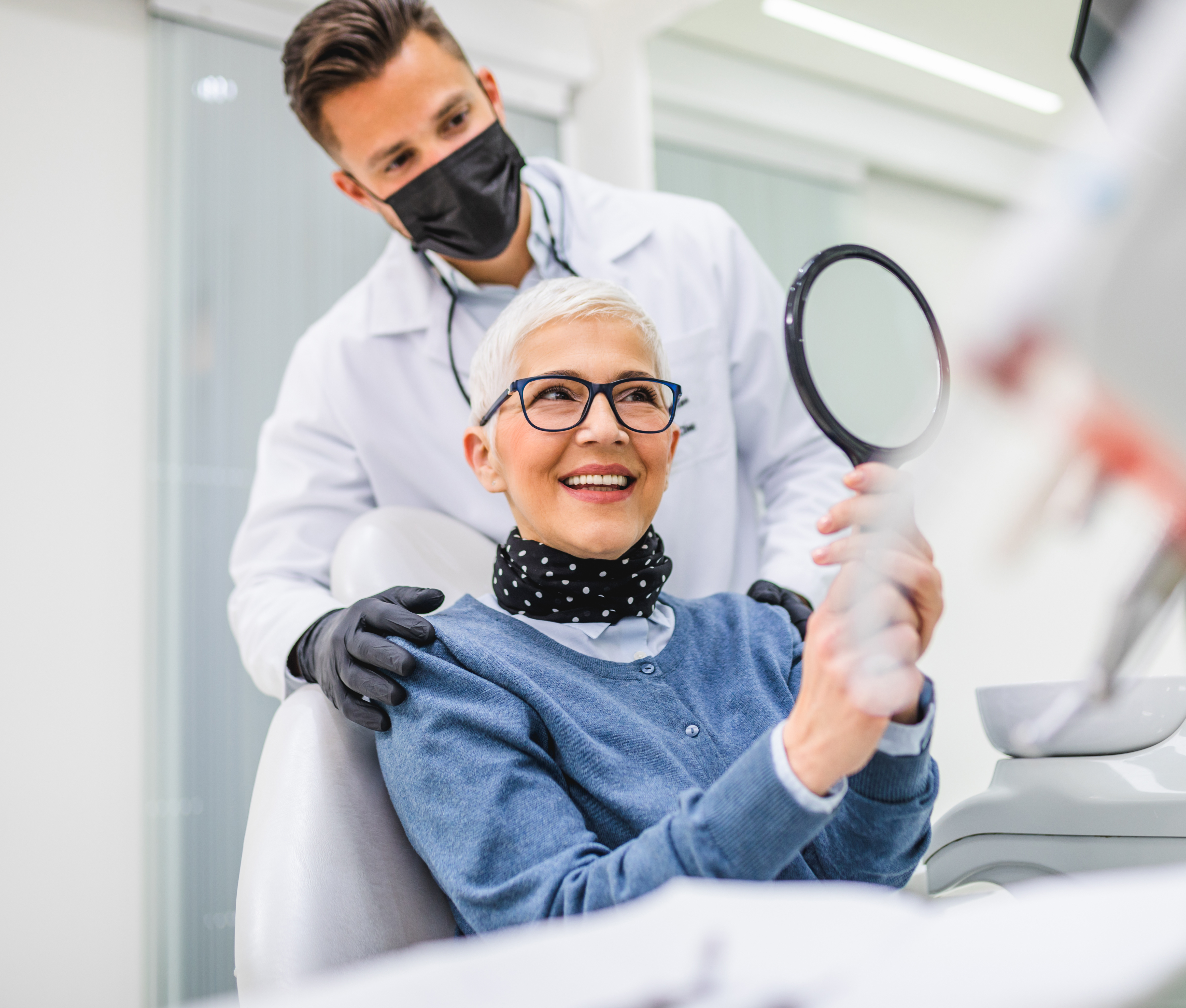 Ältere Patientin sitzt auf dem Behandlungsstuhl einer Zahnarztpraxis und lächelt in einen Handspiegel, den sie in ihren Händen hält. Hinter ihr steht der behandelnde Zahnarzt.