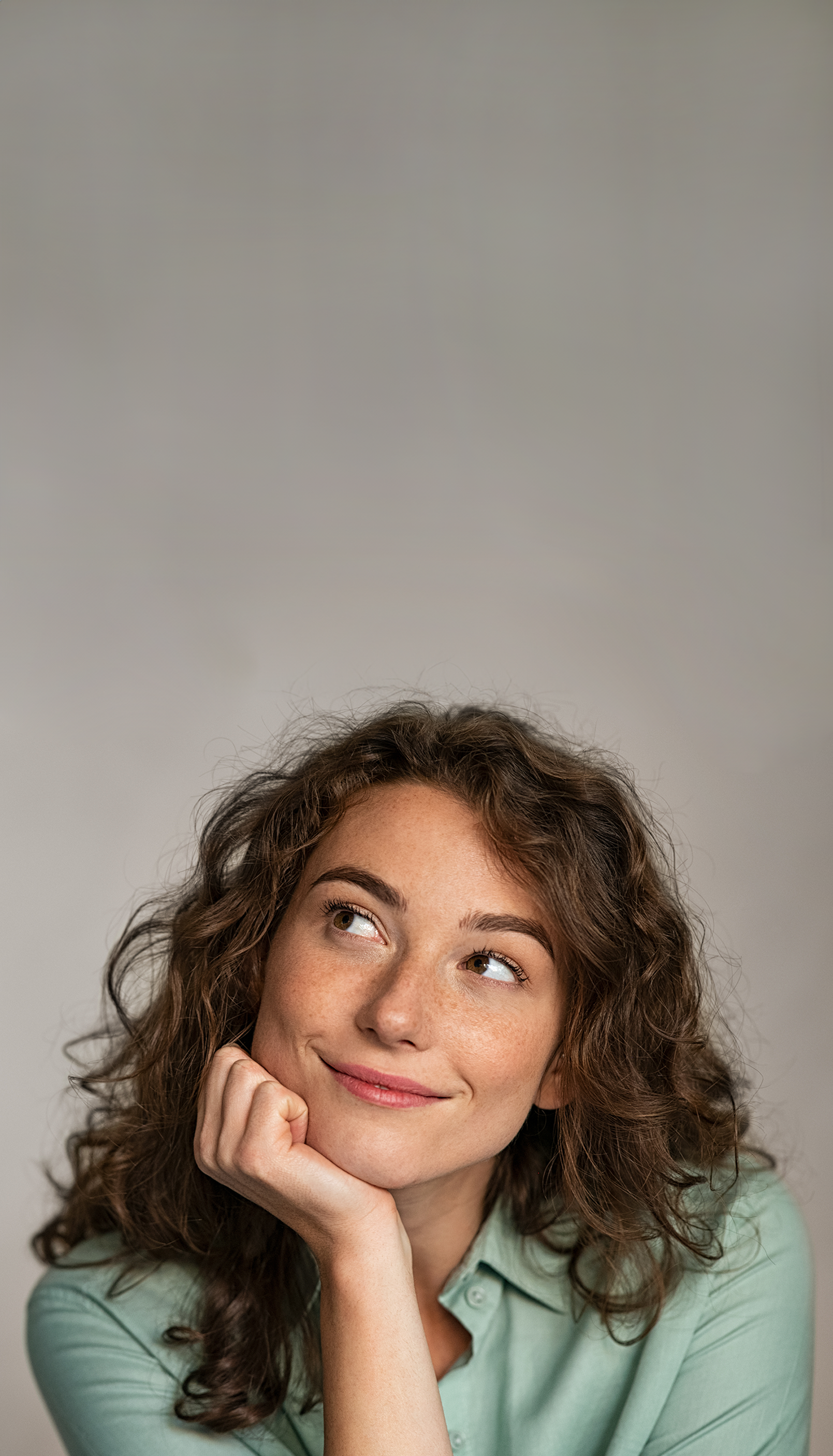Junge Frau mit braunem lockigem Haar stützt lächelnd mit der rechten Hand ihren Kopf und blickt zur linken Seite.