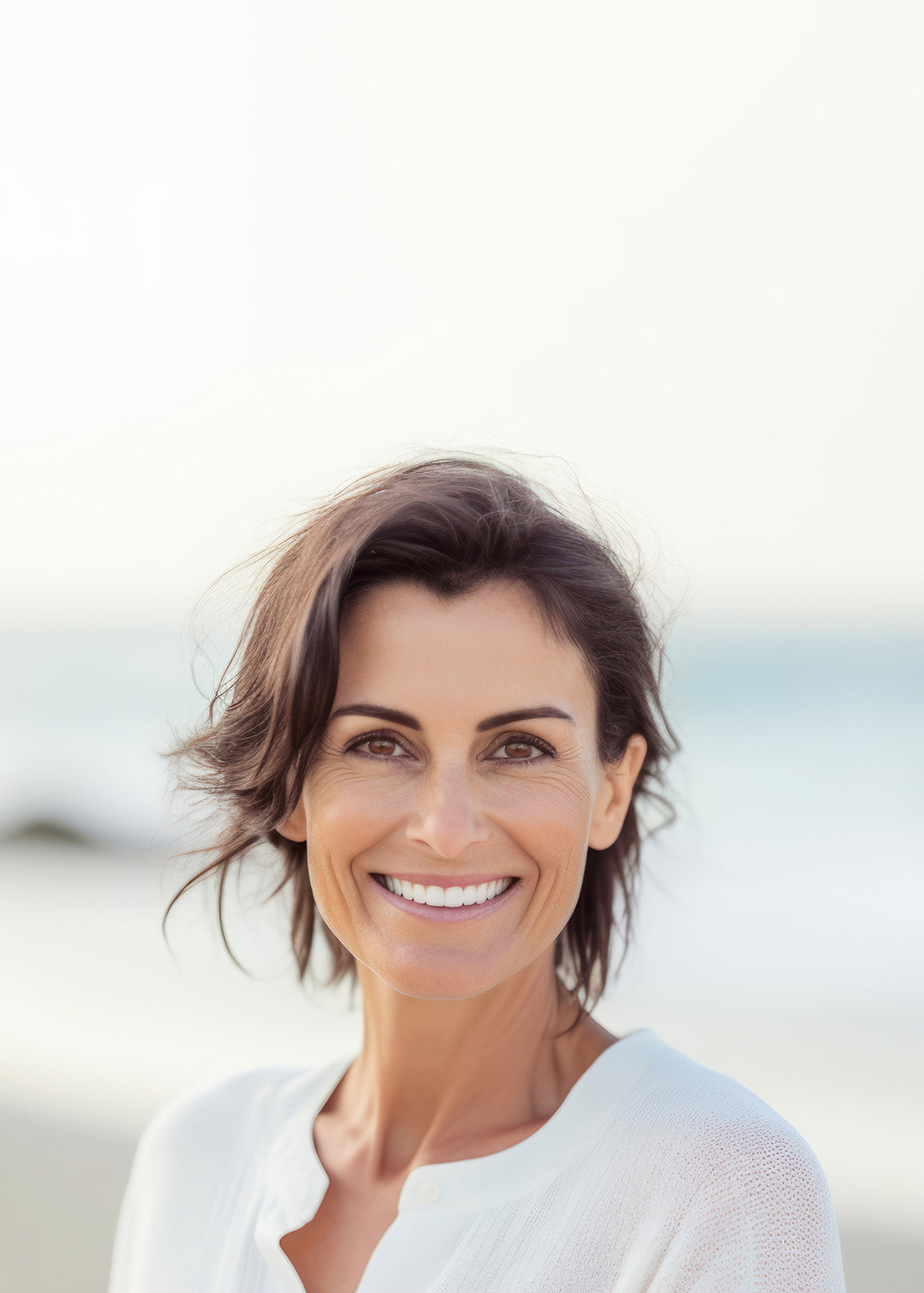 Ältere Frau mit braunen Haaren trägt eine weiße Bluse und lacht Zähne zeigend. Sie befindet sich am Strand mit Meer im Hintergrund.