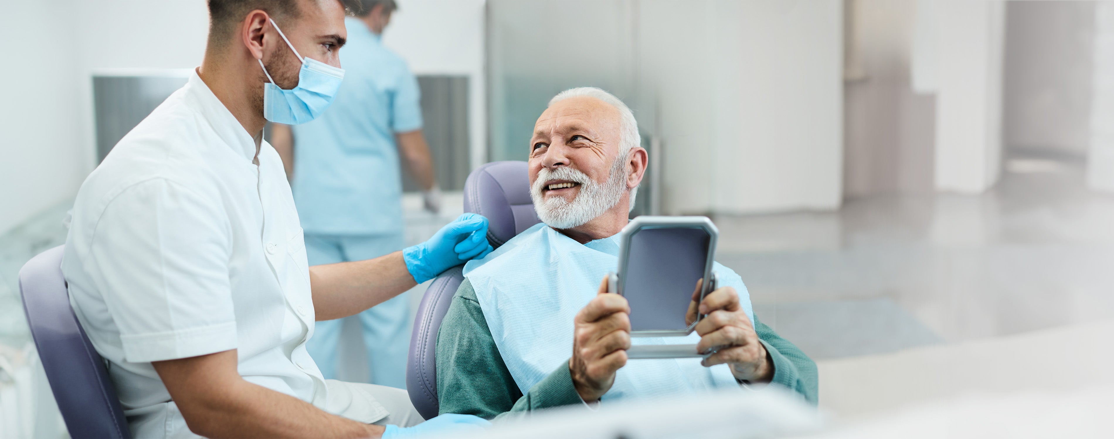 Älterer Patientin sitzt auf dem Behandlungsstuhl einer Zahnarztpraxis und hält einen Spiegel in seinen Händen. Neben ihn sitzt ein junger Zahnarzt, beide schauen sich gegenseitig an.