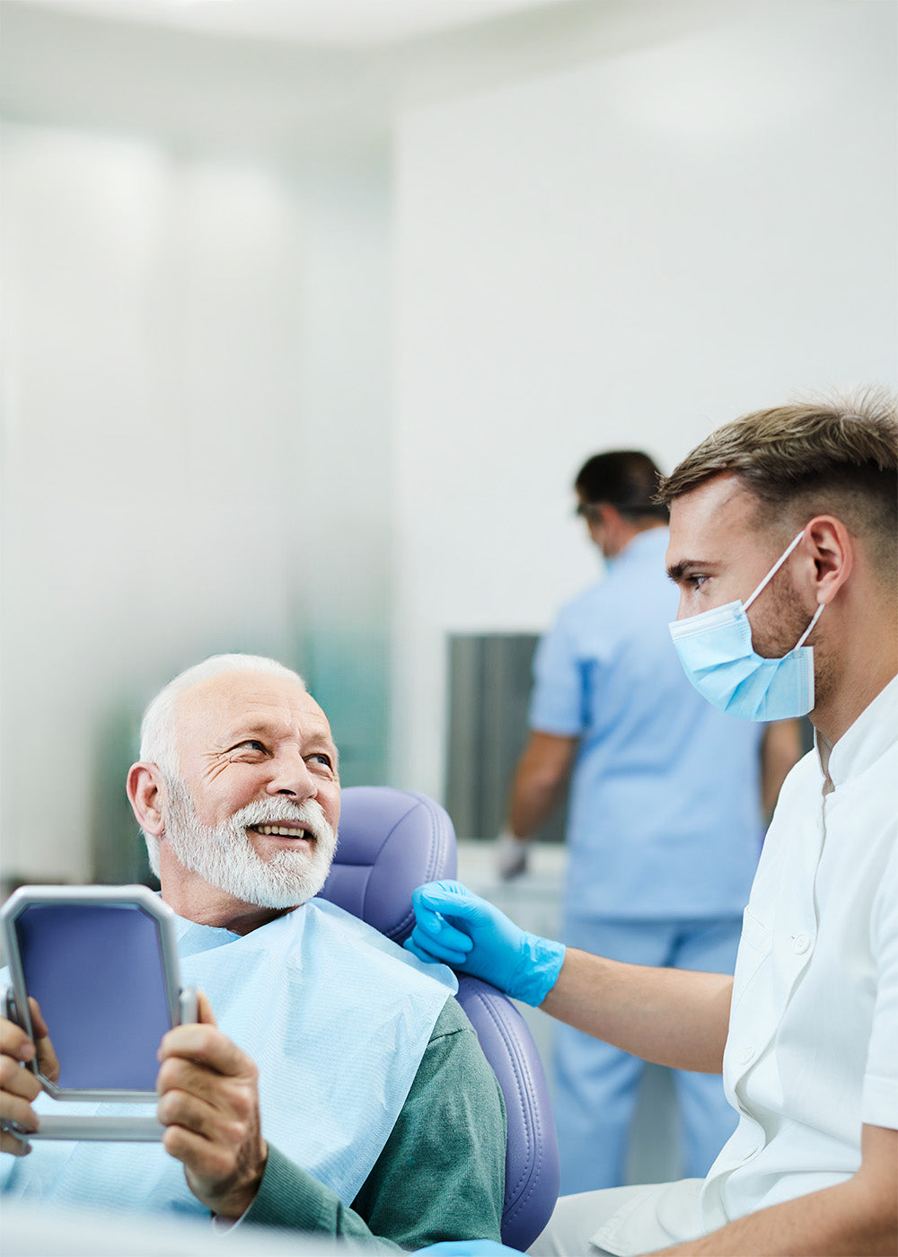 Männlicher Patient sitzt auf dem Behandlungsstuhl und hält einen Spiegel in seinen Händen. Daneben der Zahnarzt, beide Personen blicken sich an.