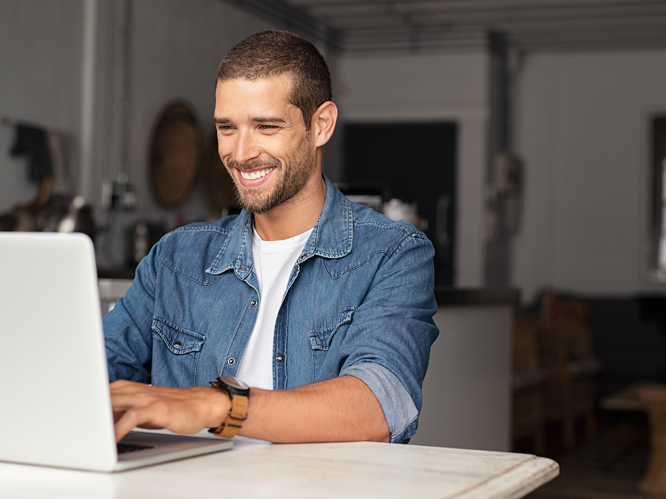 Junger Mann mit kurzen braunen Haaren trägt ein blaues Jeanshemd und sitzt lächelnd am Laptop.