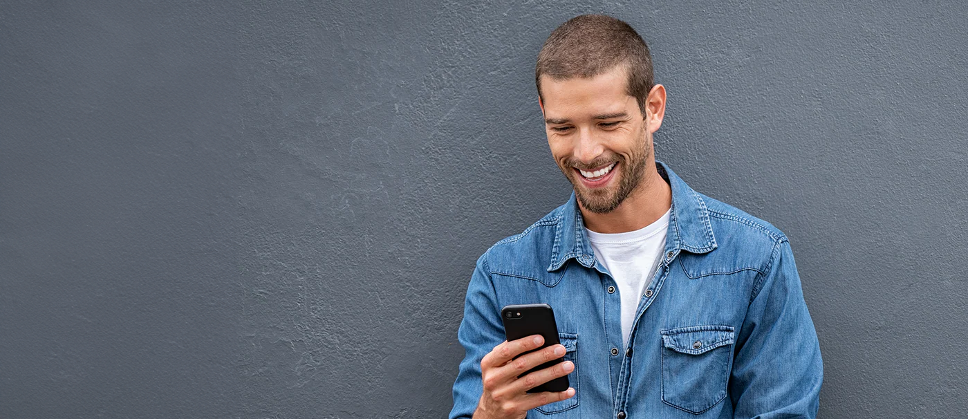 Junger Mann im blauen Jeanshemd, der lächelnd in sein Smartphone schaut.