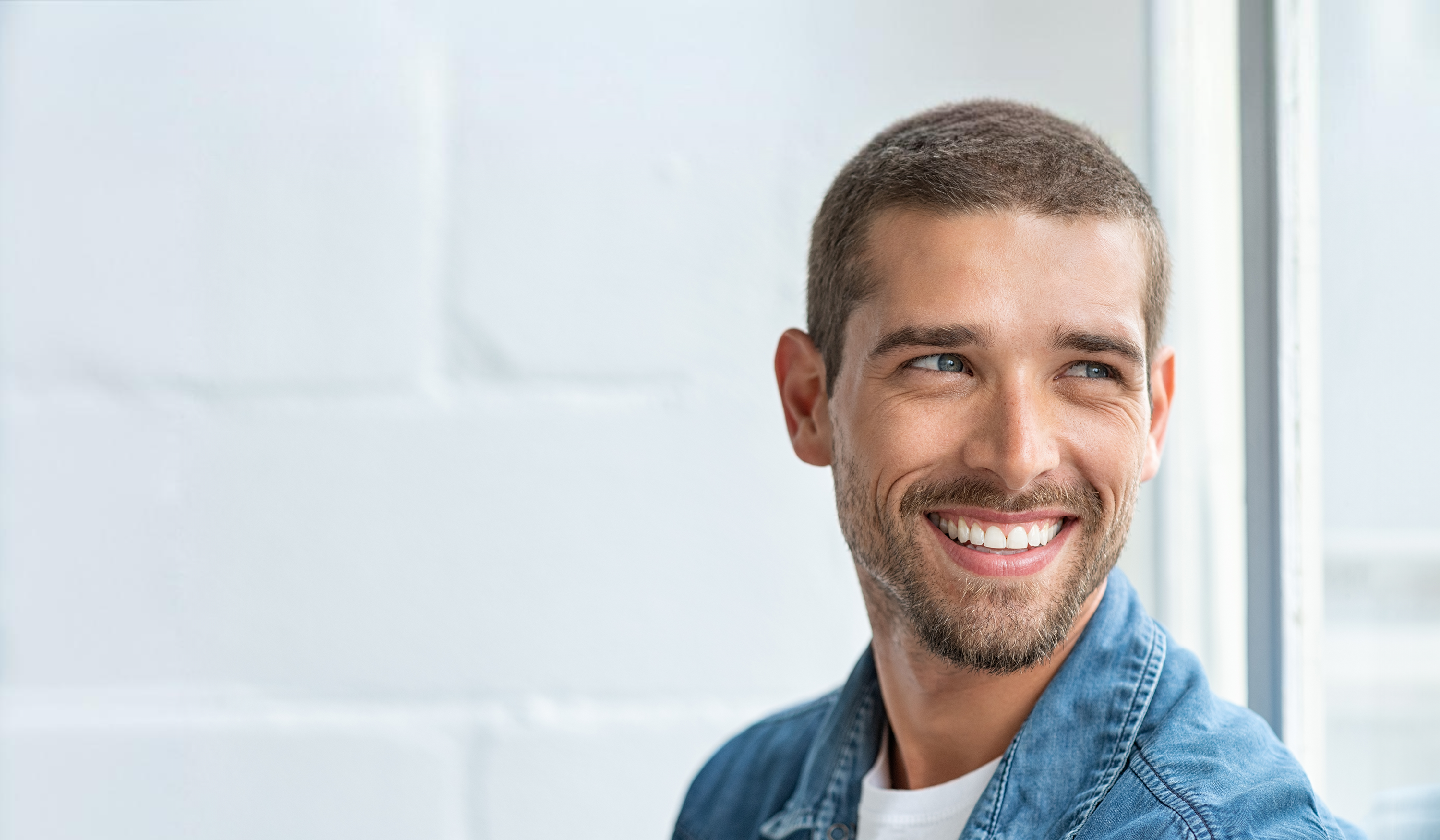 Junger Mann mit kurzen braunen Haaren und blauen Augen im blauen Jeanshemd der lächelnd zur Seite blickt.