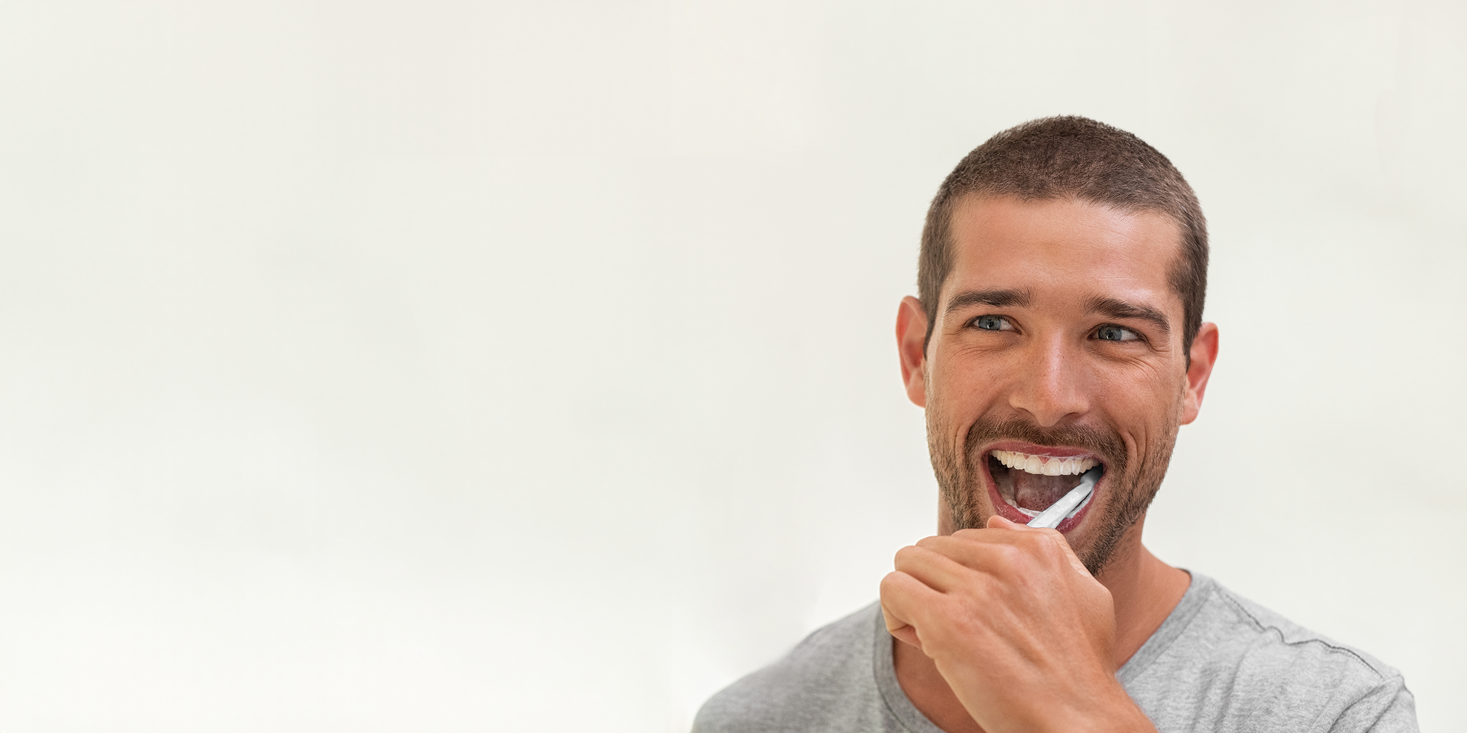 Junger Mann mit kurzen braunen Haaren putzt seine Zähne mit einer weißen Hand-Zahnbürste.