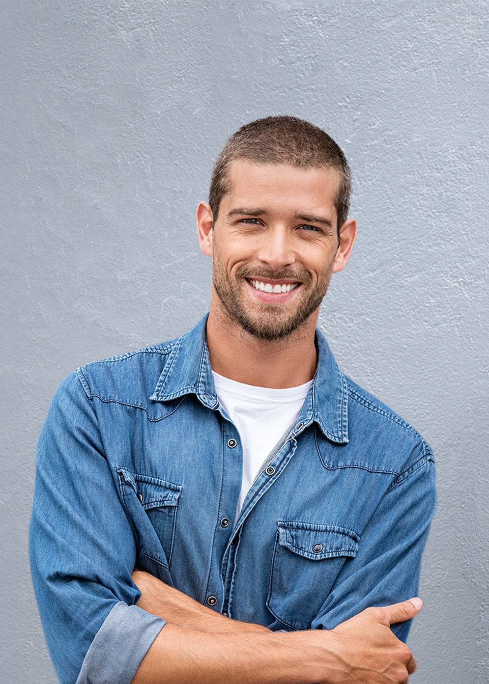Junger Mann mit kurzen braunen Haaren, im blauen Jeanshemd, lehnt an einer dunkelgrauen Hauswand. Er hat dabei die Arme verschränkt und lächelt.