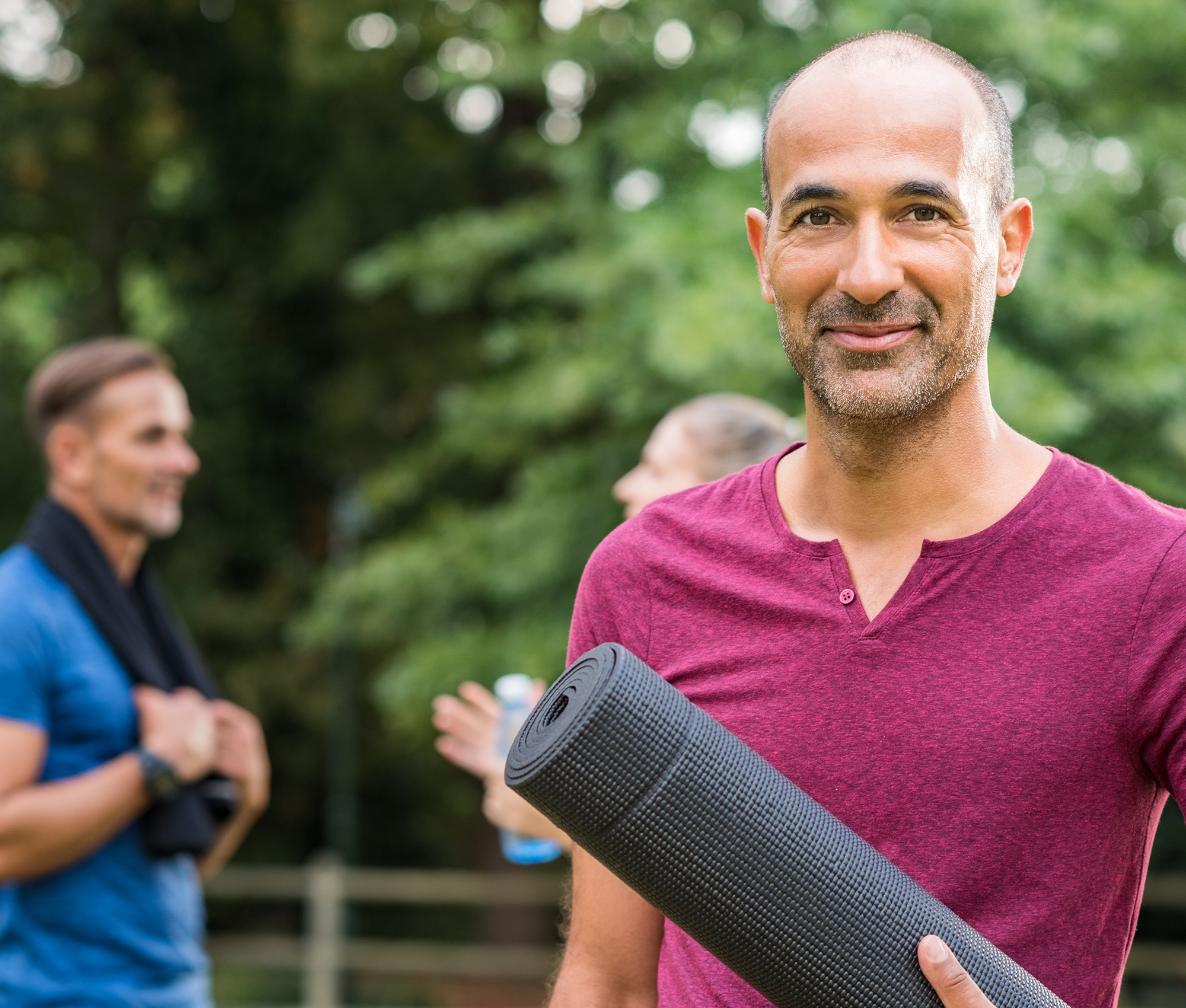 Mann im roten T-Shirt und einer schwarzen Fitness-Matte in der Hand blickt lächelnd in die Kamera. Im Hintergrund sind weitere Personen in Sportkleidung erkennbar.