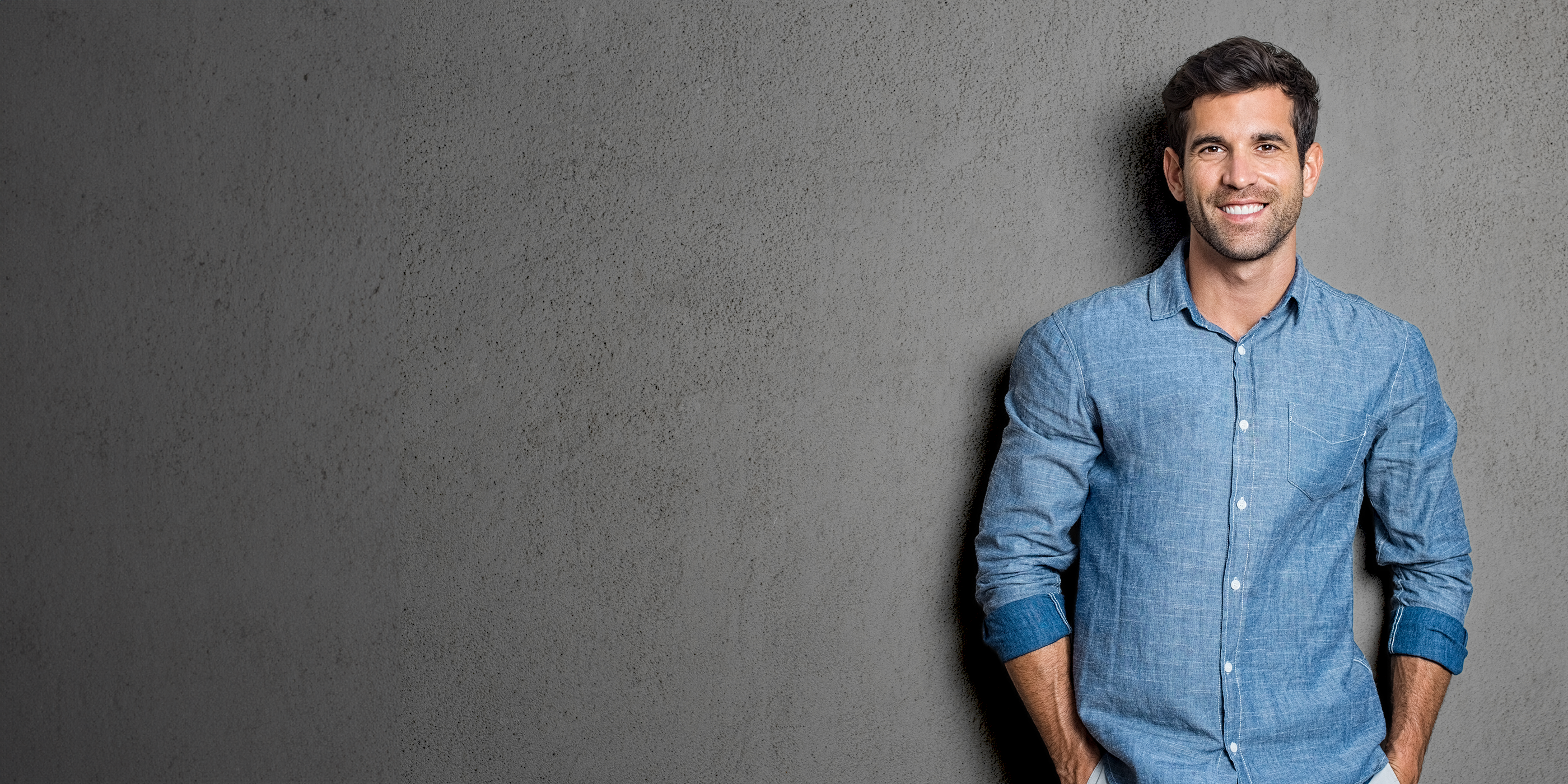Junger Mann mit kurzen braunen Haaren, im blauen Jeanshemd, lehnt an einer grauen Hauswand. Er hat dabei die Arme in seinen Hosentaschen.