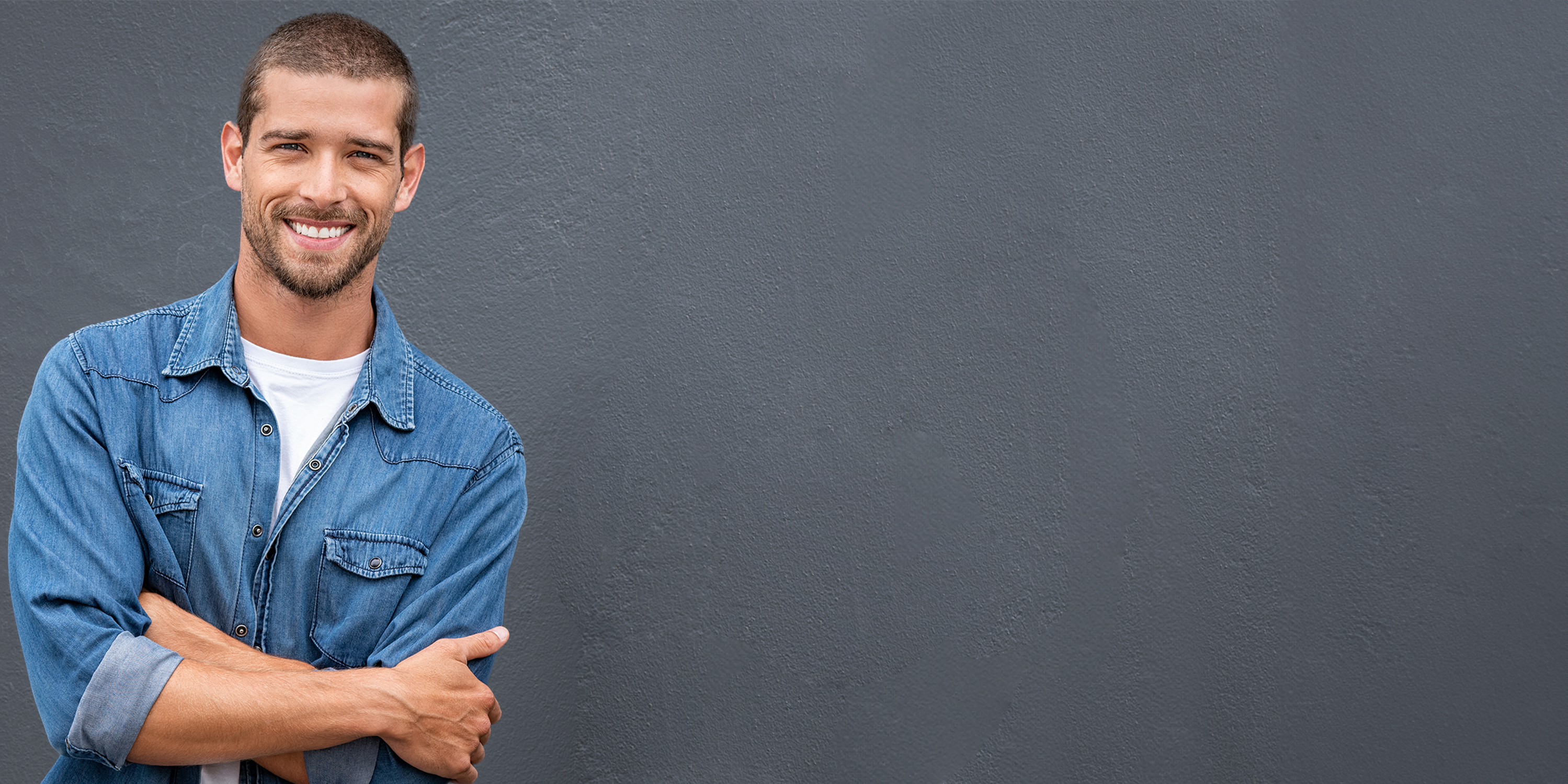 Junger Mann mit kurzen braunen Haaren, im blauen Jeanshemd, lehnt an einer grauen Hauswand. Er hat dabei die Arme verschränkt und lächelt.