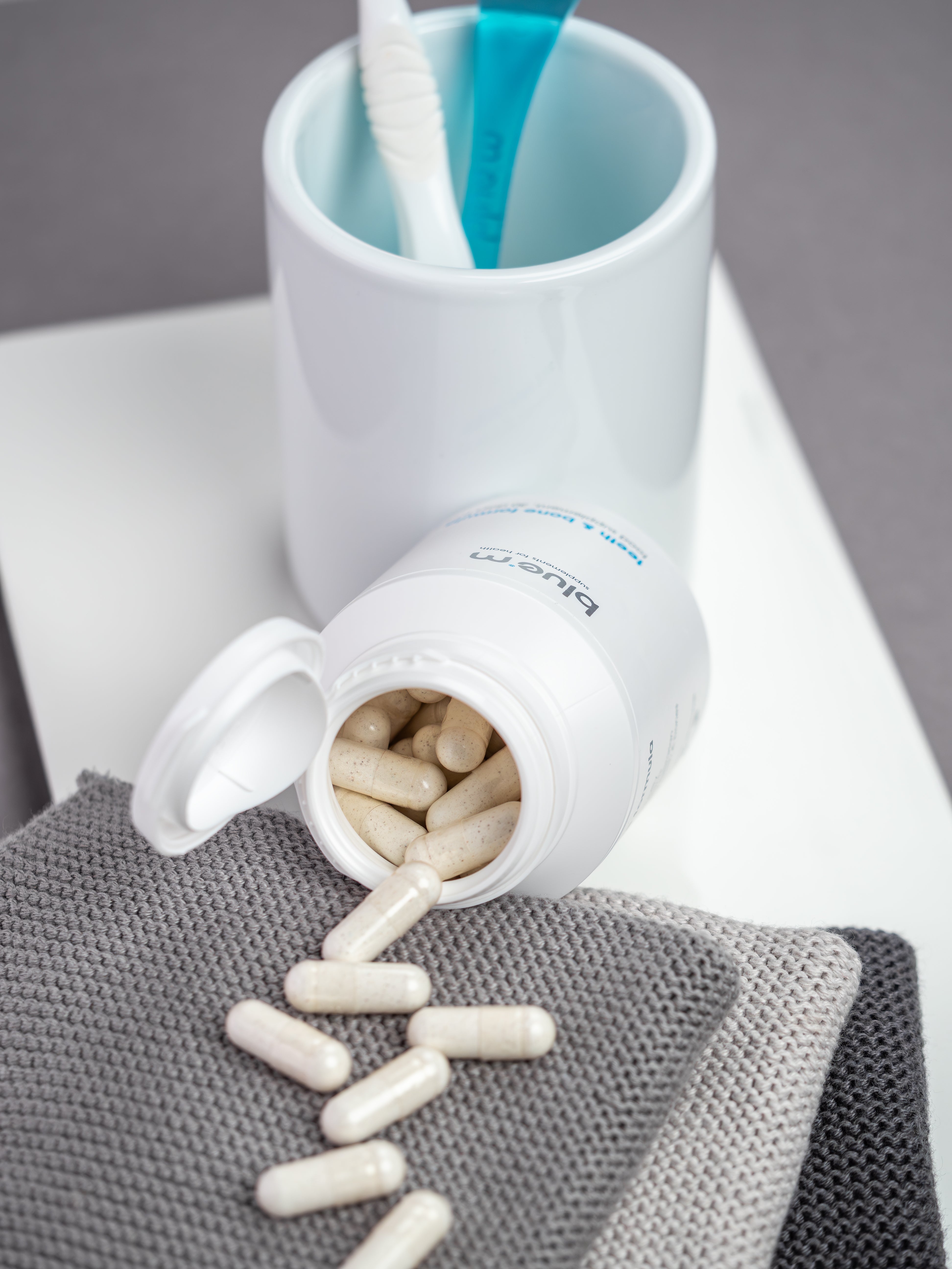 Geöffnete Dose von Nahrungsergänzung-Kapseln für Knochen und Zahn, davor liegen die Kapseln verstreut auf einem grauen Handtuch, im Hintergrund ist ein Zahnputzbecher mit Zahnbürste und Zungenreiniger erkennbar.