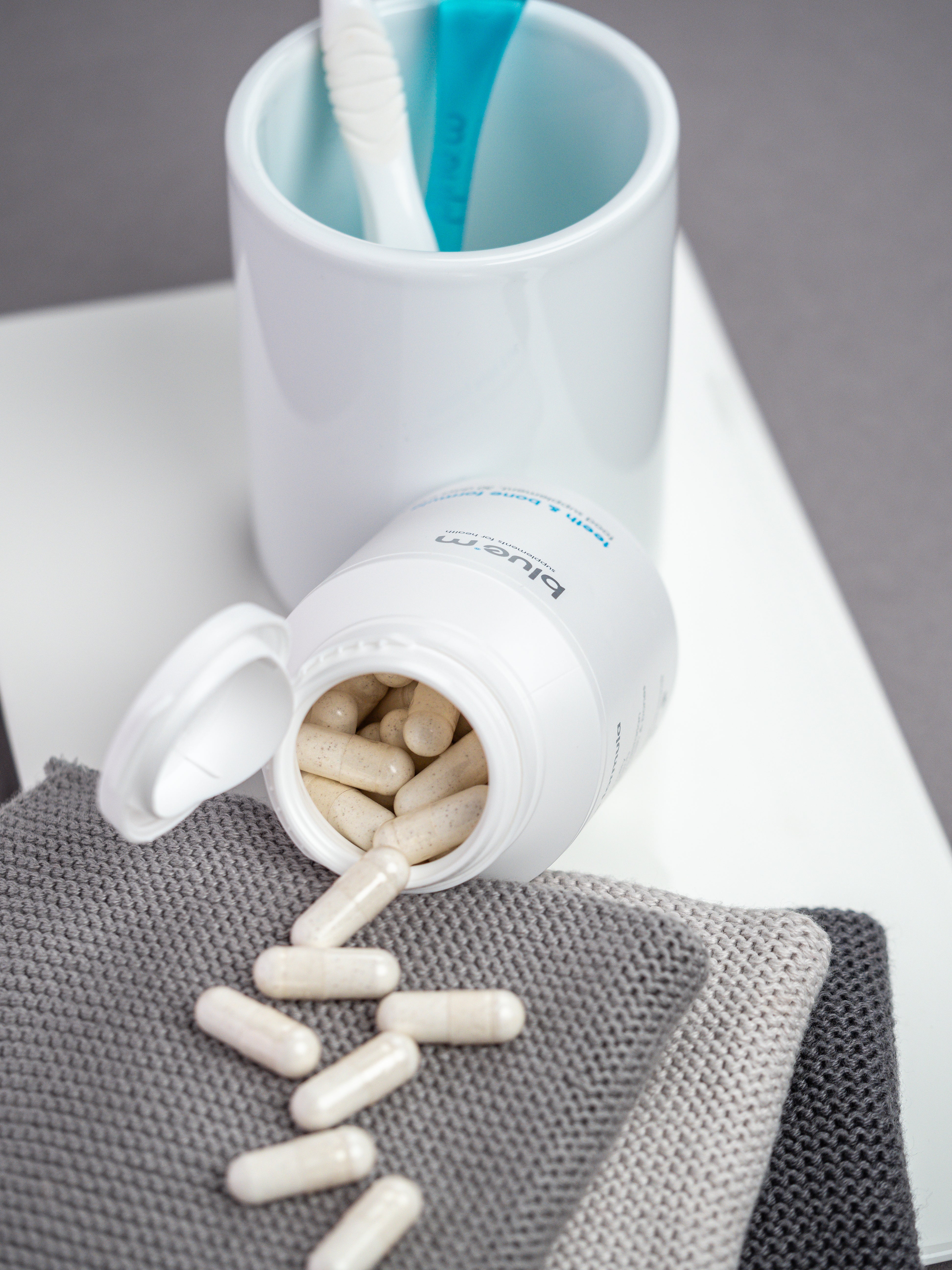 Geöffnete Dose von Nahrungsergänzung-Kapseln für Knochen und Zahn, vor der Dose liegen die Kapseln verstreut auf dem Untergrund. Im Hintergrund ist ein Zahnputzbecher mit Zungenreiniger und Zahnbürste erkennbar.