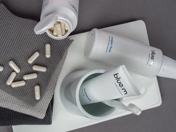 Nahrungsmittelergänzung in Form von Kapseln liegen verstreut auf einem Stapel an Handtüchern, rechts daneben befindet sich eine Flasche Sauerstoff-Fluid und in einem Zahnputzbecher Zahnpasta und eine Zahnbürste.