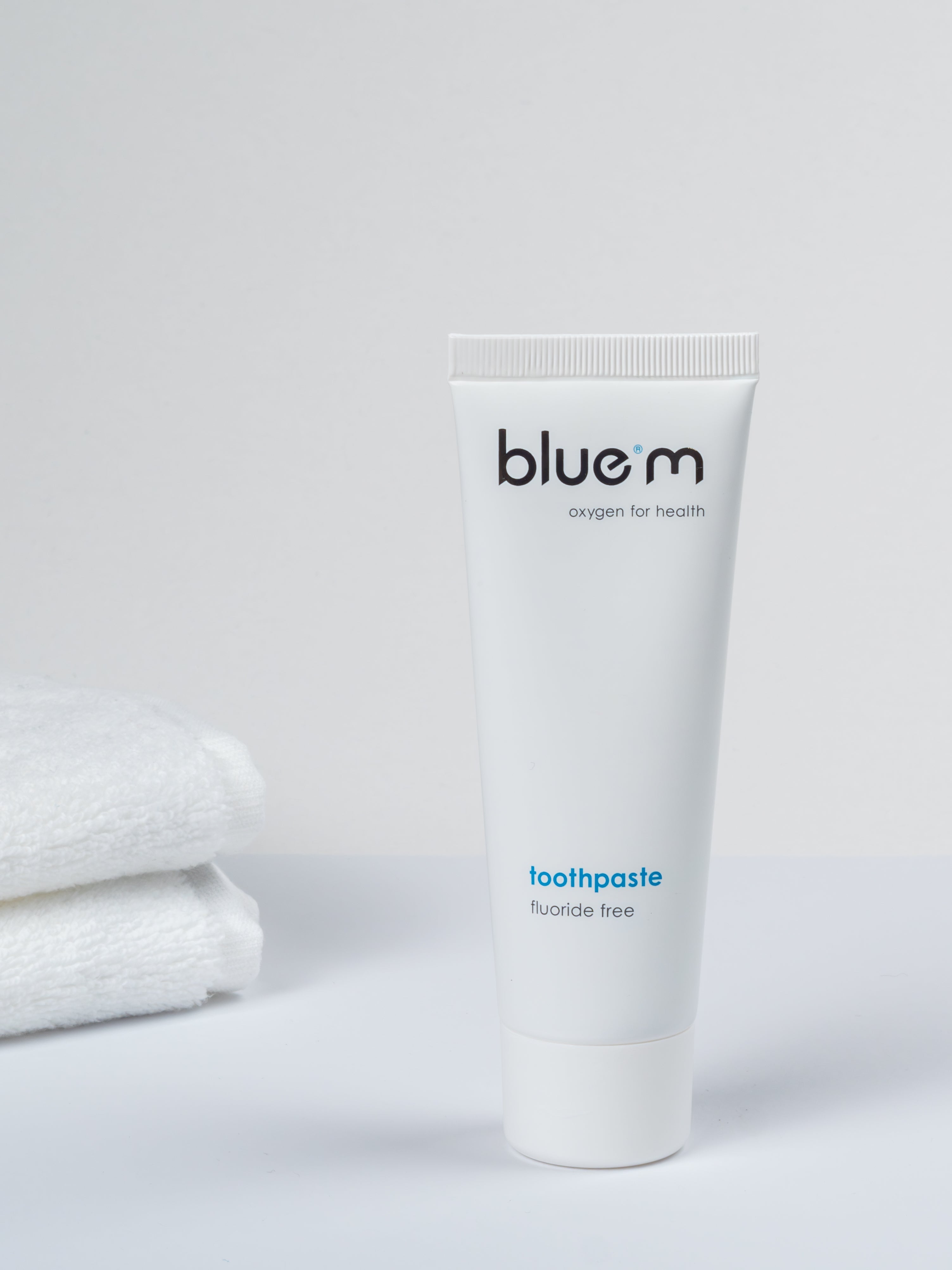 Eine Tube Zahngel ohne Fluorid auf weißem Untergrund stehend, im Hintergrund ist ein Stapel Handtücher erkennbar.