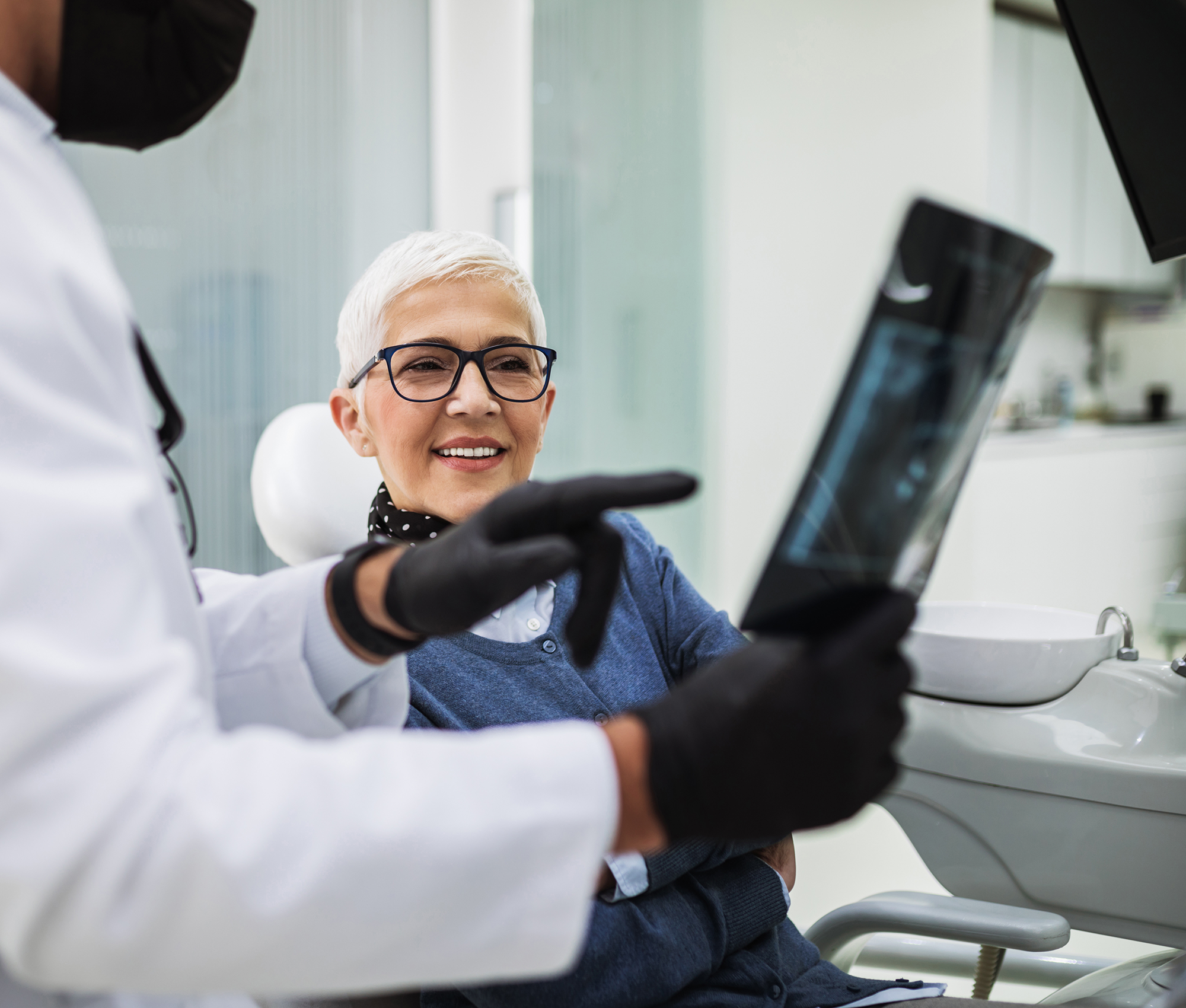 Zahnarzt erklärt mittels eines Röntgenbildes einer Patientin mit kurzen weißen Haaren und schwarzer Brille den Behandlungsplan.