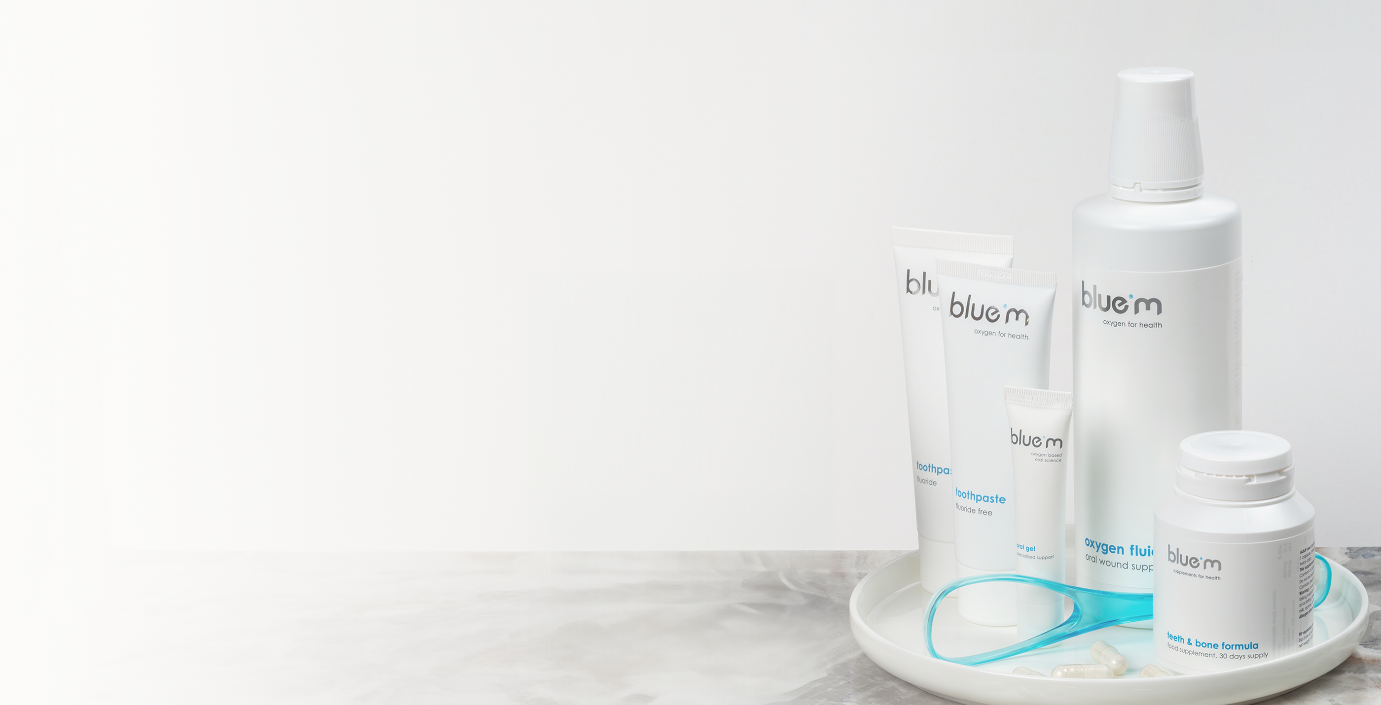 Pflegeset für eine optimale Mundpflege bestehend aus Nahrungsmittelergänzungs-Kapseln, Wundgel, Zahngel, Zungenreiniger und Sauerstoff-Fluid arrangiert auf einem weißen Tabelett dabenen eine weiße Orchidee in einer kleinen Vase.