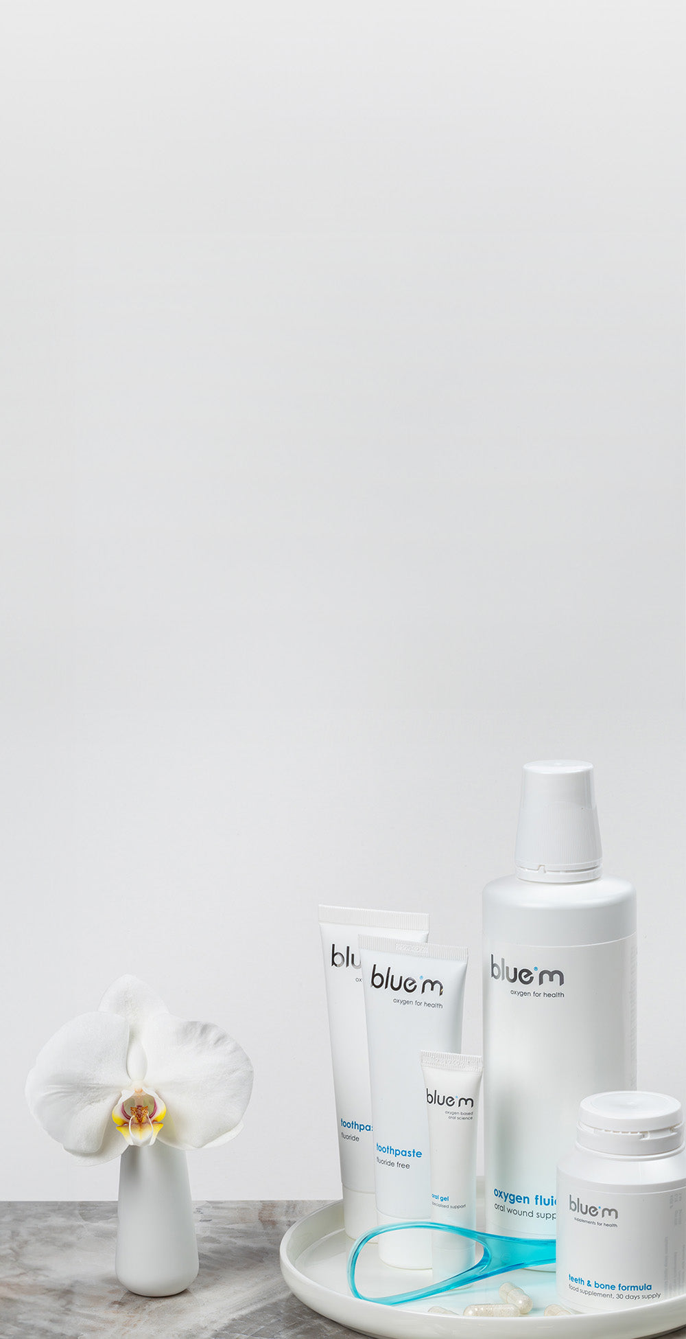 Pflegeset für eine optimale Mundpflege bestehend aus Nahrungsmittelergänzungs-Kapseln, Wundgel, Zahngel, Zungenreiniger und Sauerstoff-Fluid arrangiert auf einem weißen Tabelett dabenen eine weiße Orchidee in einer kleinen Vase.