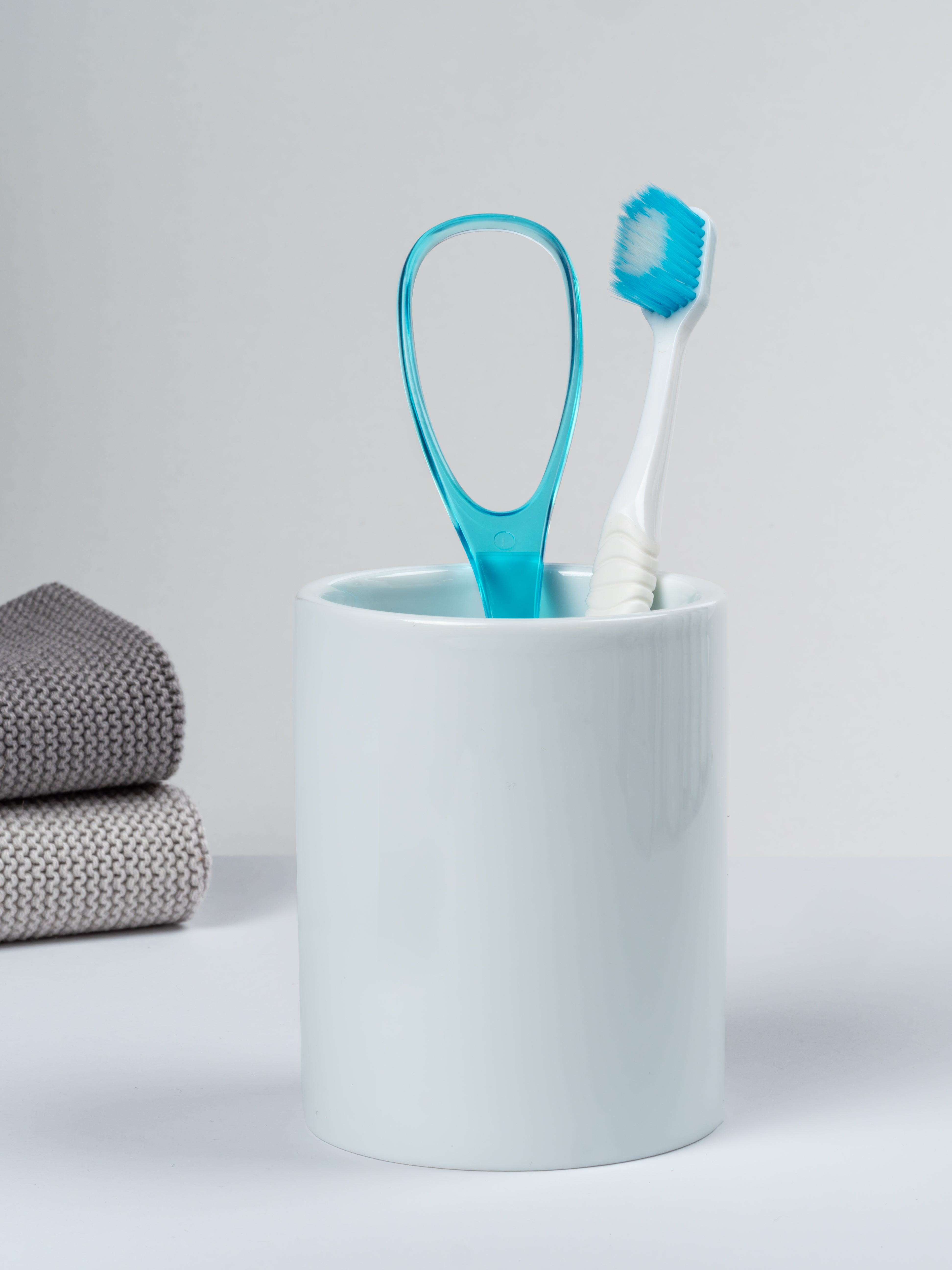 Komfort-Zahnbürste mit hellblauen Bürsten und hellblauer Zungenreiniger in einem Zahnputzbecher stehend.