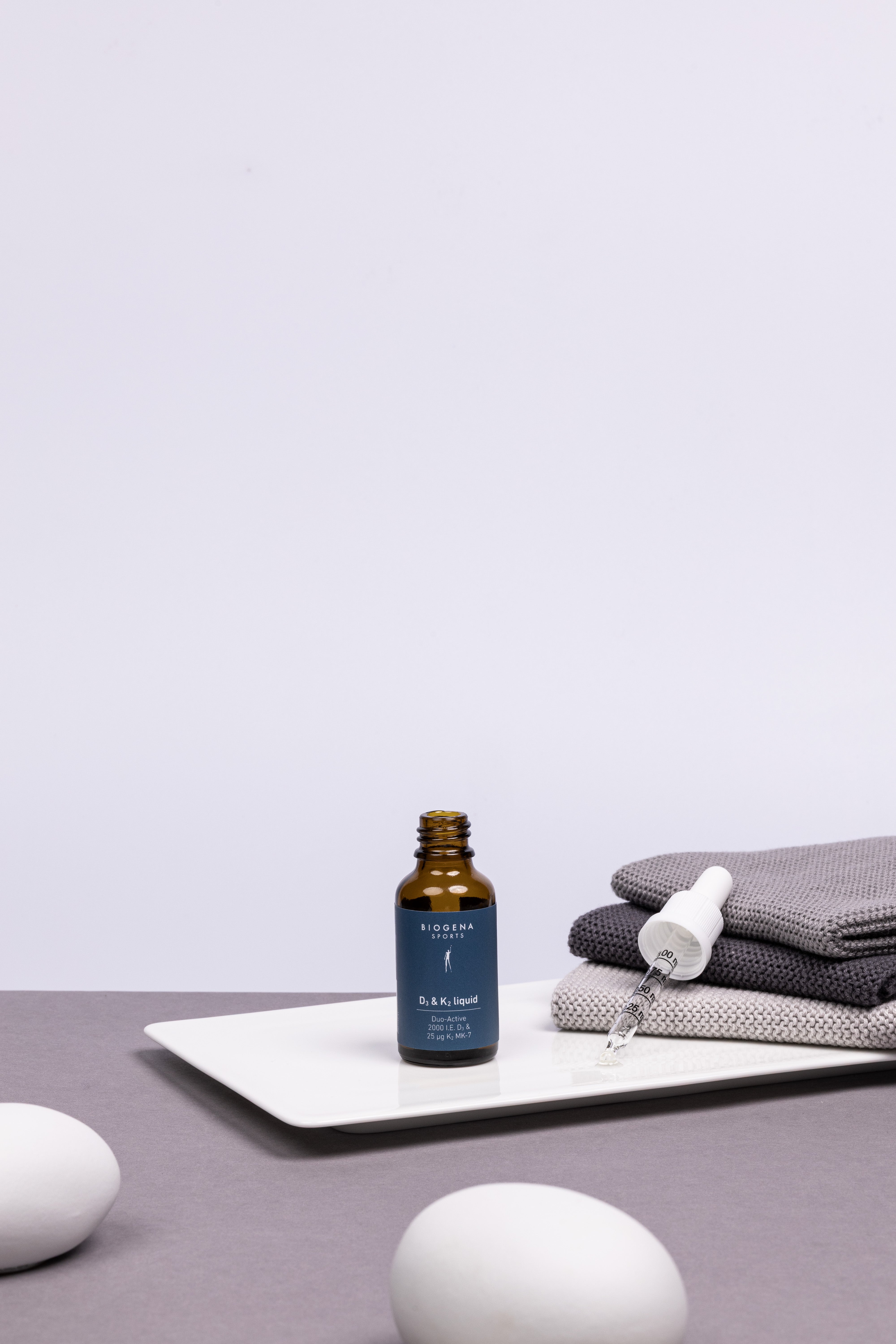 Geöffnete Flasche BIOGENA SPORTS D3 & K2 liquid Flasche stehend, daneben der Flaschendeckel mit Pipette auf einem Stapel grauer Handtücher liegend.