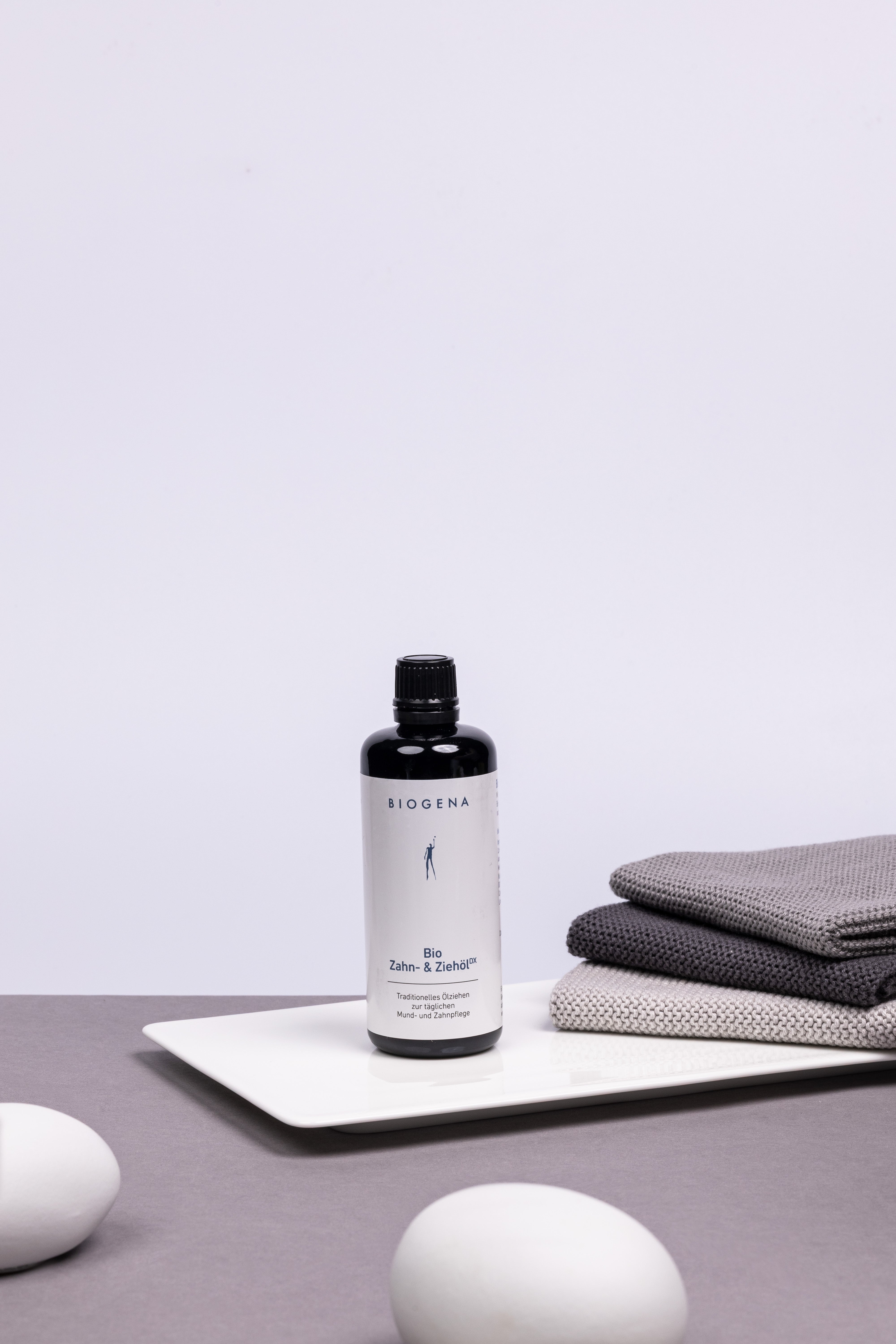 BIOGENA Zahn- und Ziehöl Flasche steht auf einem weißen Tablett, rechts daneben liegt Stapel grauer Handtücher .