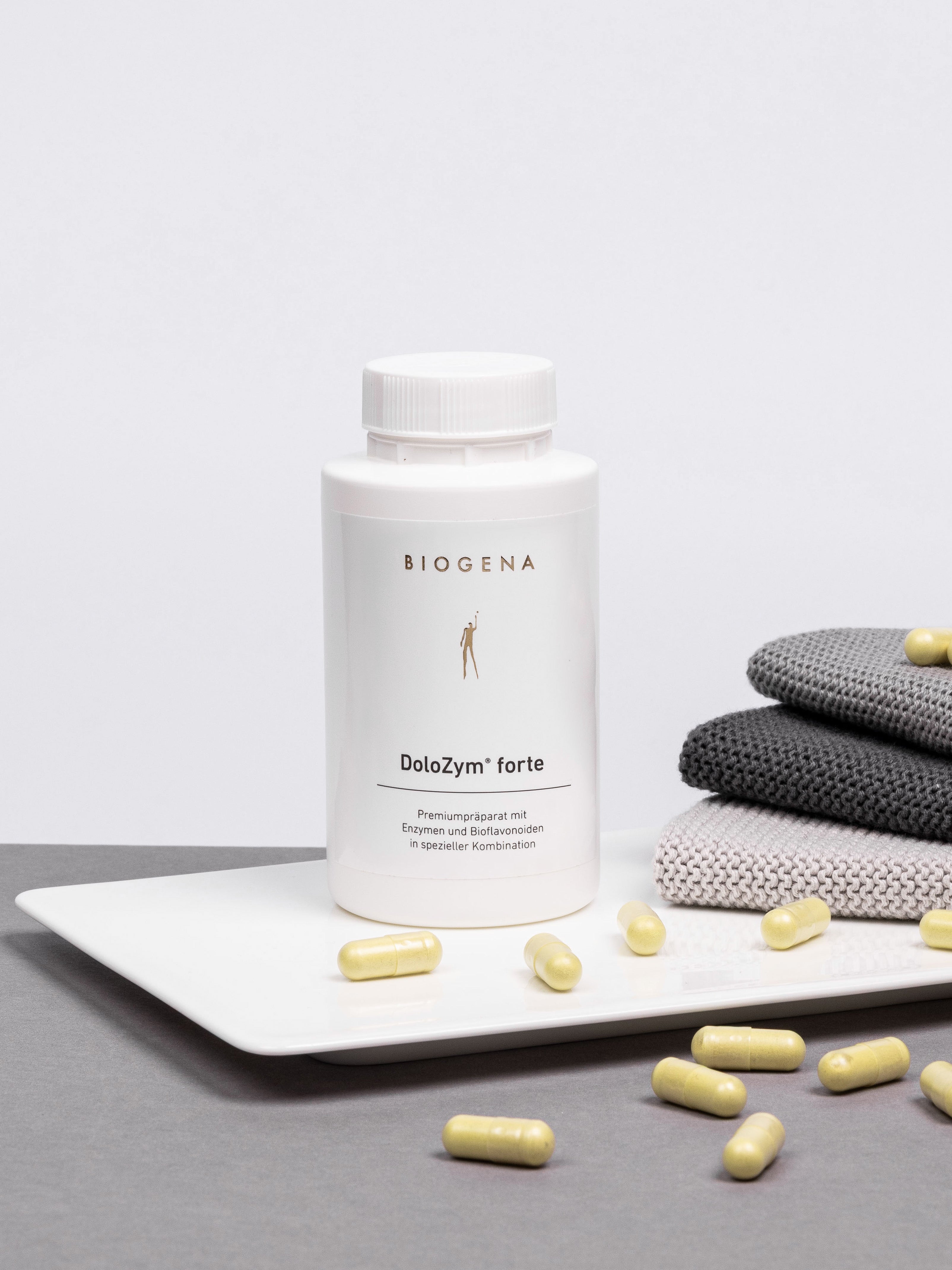 Verpackungsdose von Biogena DoloZym forte steht auf einem weißen Tablett umgeben von gelben Kapseln, rechts daneben ist ein Stapel grauer Handtücher erkennbar.