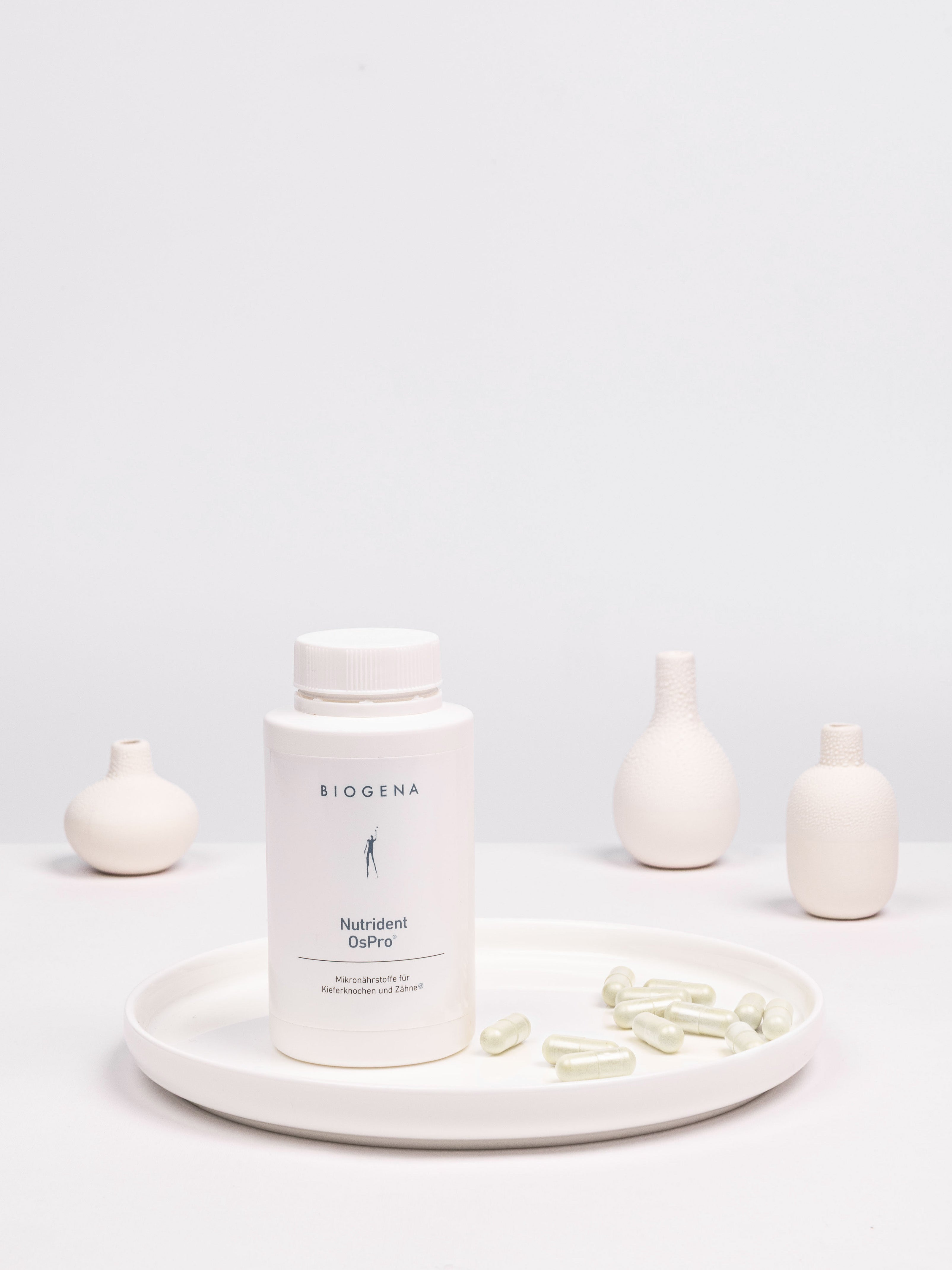 Geschlossene Verpackung von Biogena Nutrident OsPro steht auf einem weißen Tablett umgeben von weißen Kapseln.