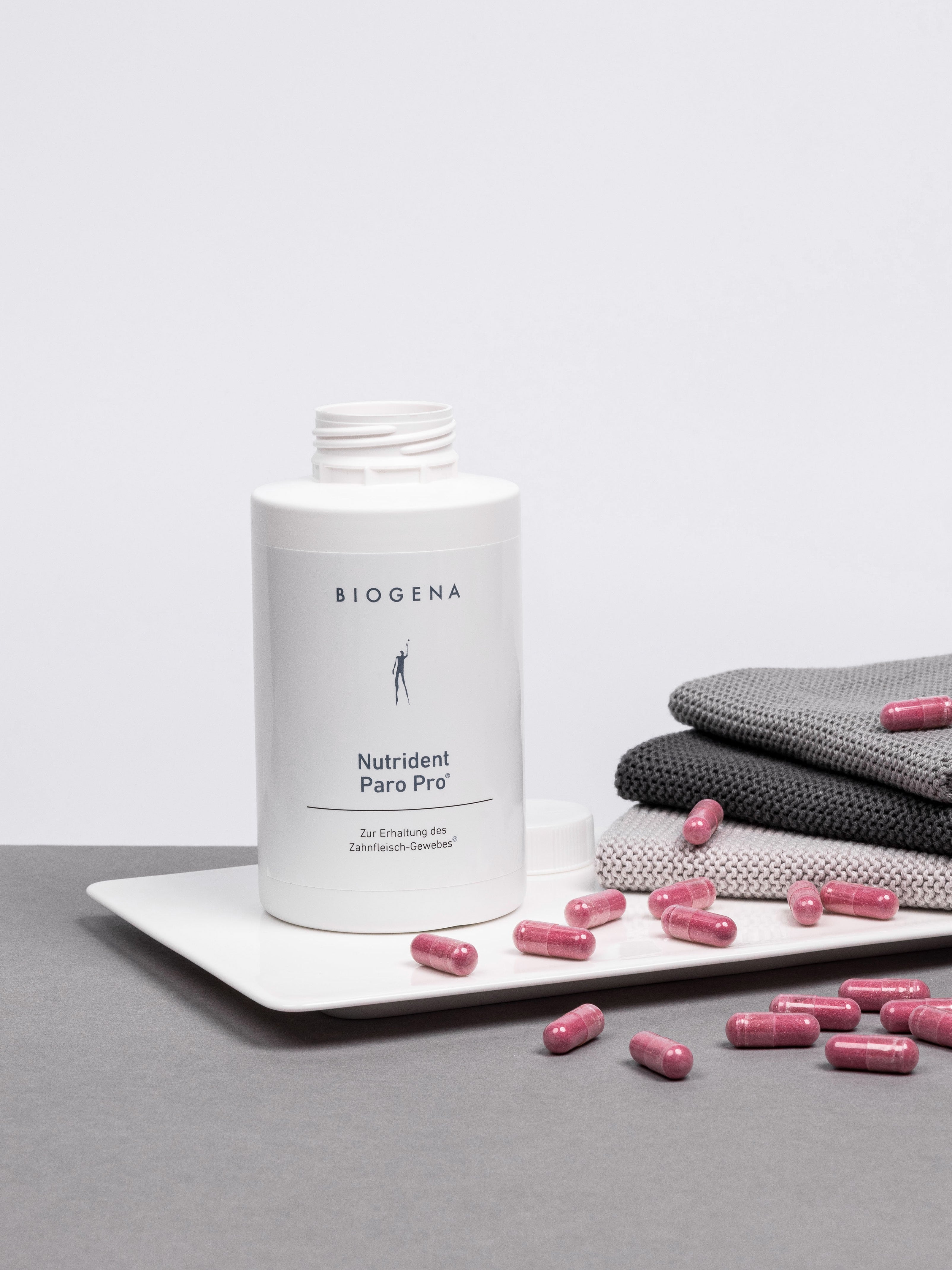 Geöffnete Verpackung von Nutrident Paro Pro steht auf einem weißen Tablett umgeben von roten Kapseln, rechts daneben ist ein Stapel grauer Handtücher erkennbar.