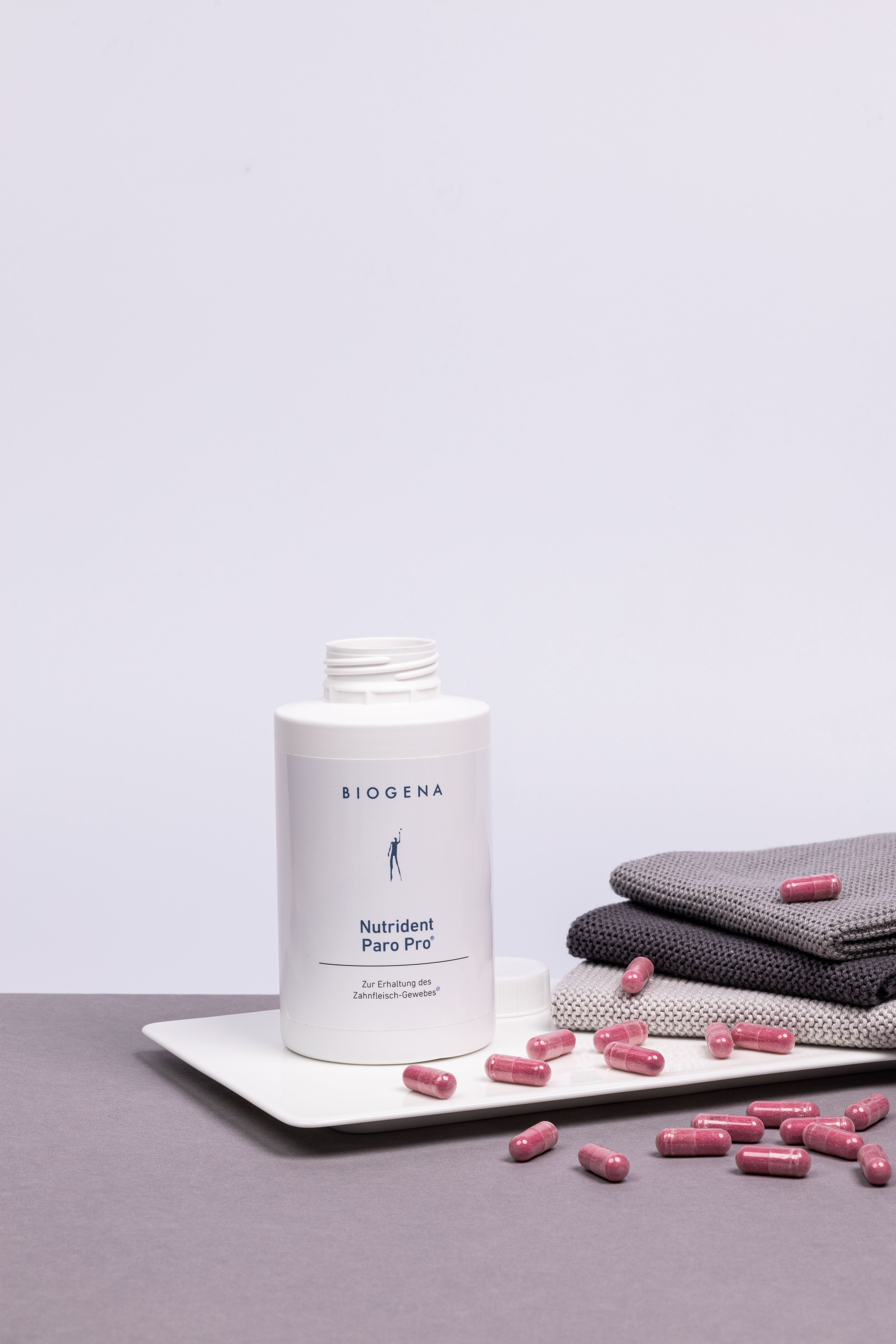 Geöffnete Verpackung von Nutrident Paro Pro steht auf einem weißen Tablett umgeben von roten Kapseln, rechts daneben ist ein Stapel grauer Handtücher erkennbar.